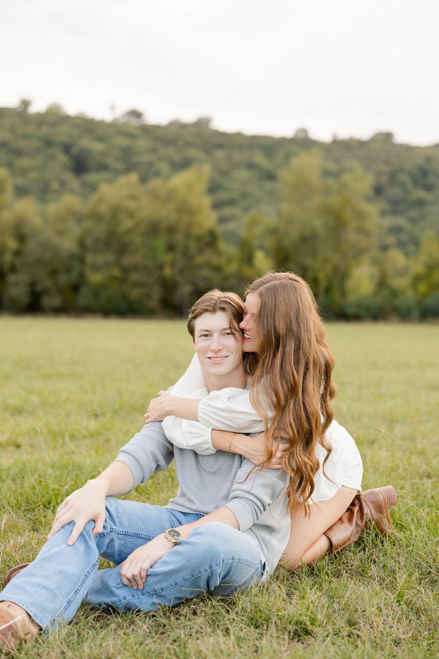 Couple lying together on grass in a serene meadow with trees and hills in the distance.