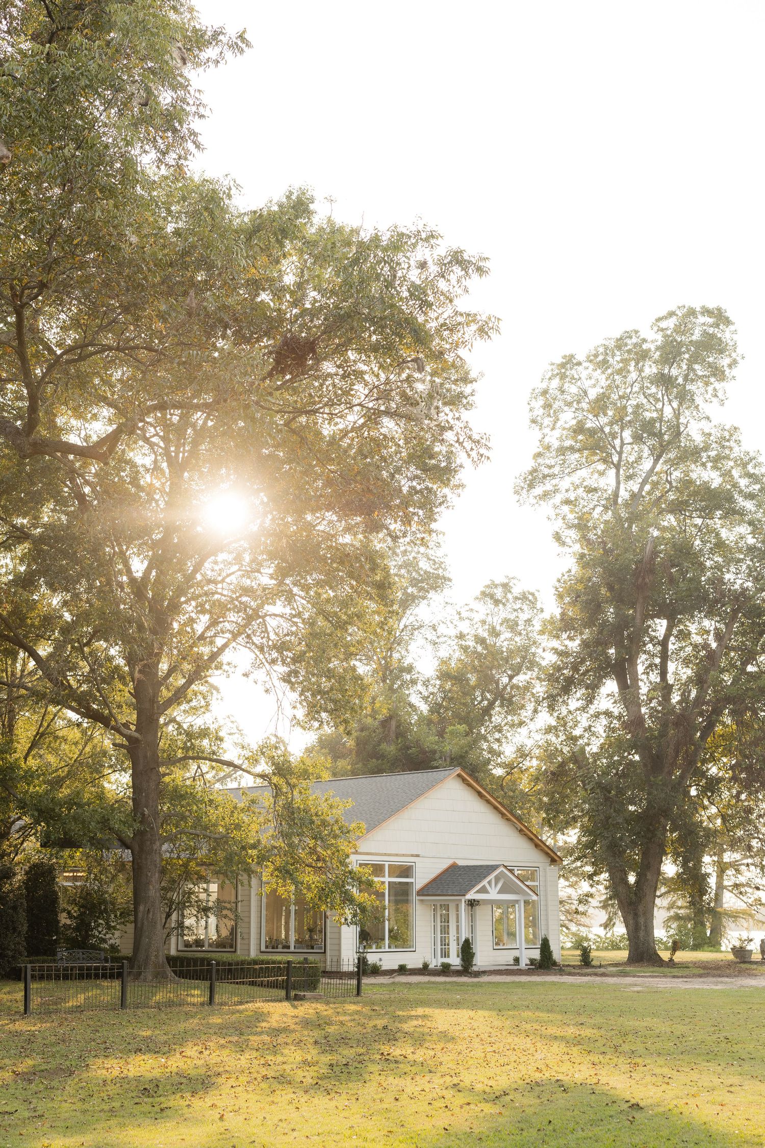 White farmhouse-style buildings nestled among trees at sunset with golden sunlight filtering through branches.