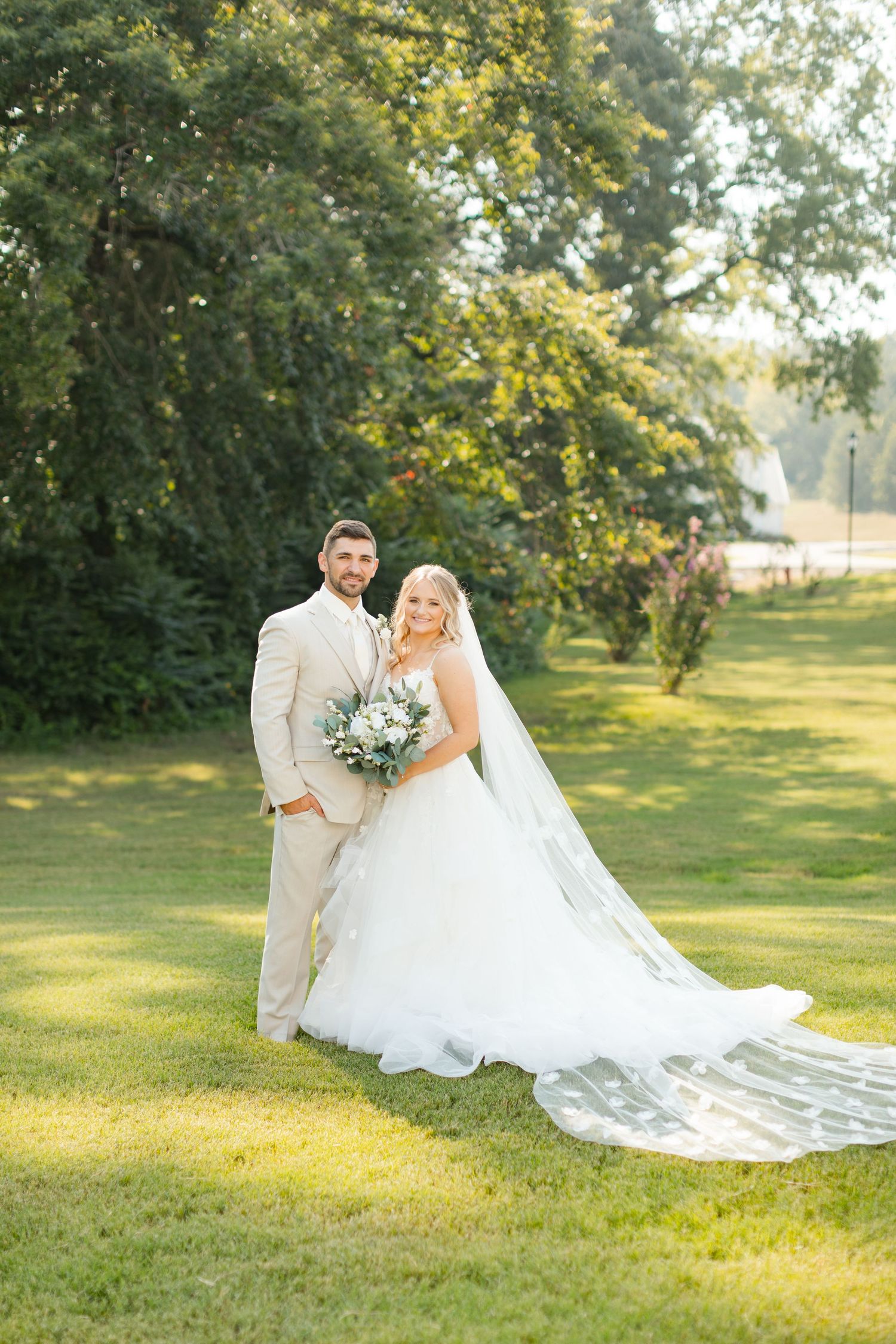 Newlyweds pose together on lush grass with a dramatic cathedral-length wedding veil trailing behind.