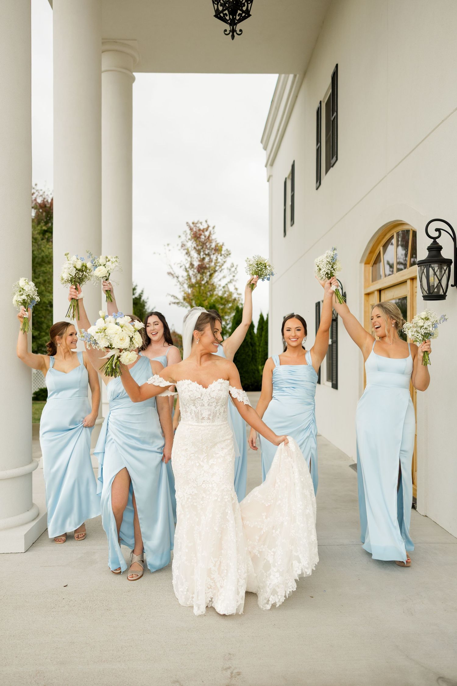 Wedding party in light blue bridesmaid dresses and white bridal gowns pose playfully along a columned porch.
