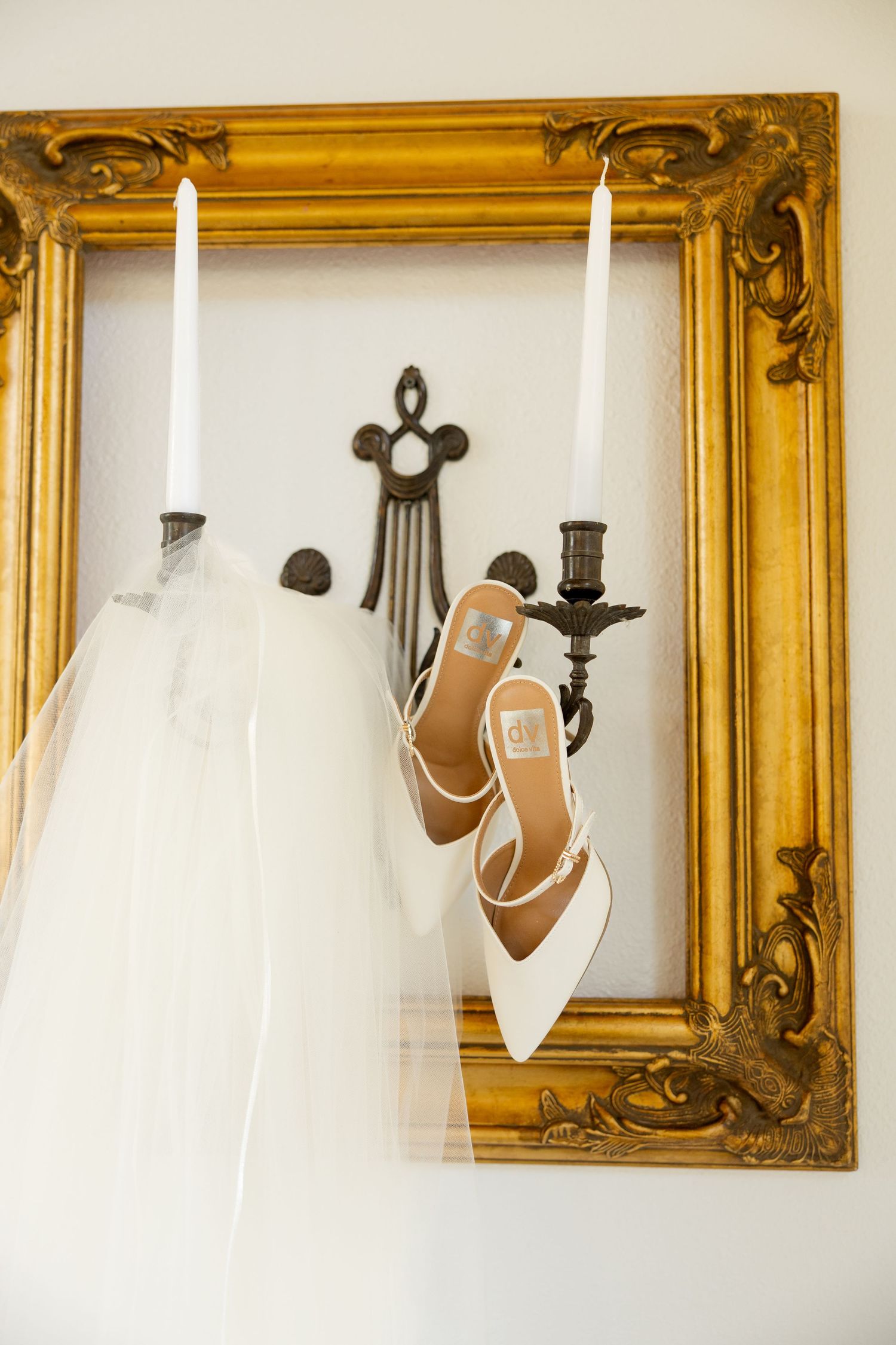 White bridal shoes and veil displayed against ornate gold-framed mirrors on a light colored wall.