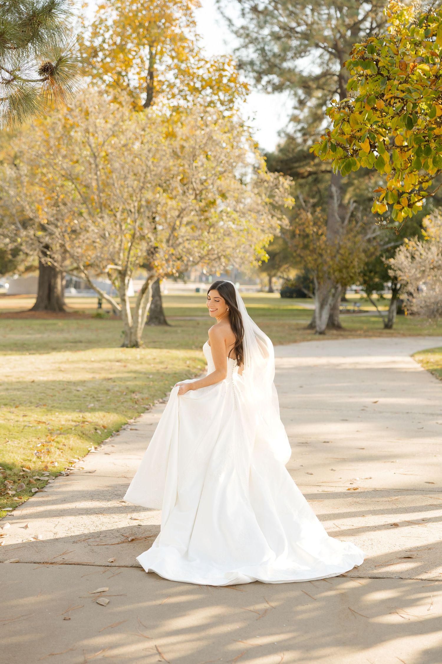 A bride in a flowing white wedding gown stands on a path surrounded by autumn trees with golden foliage.