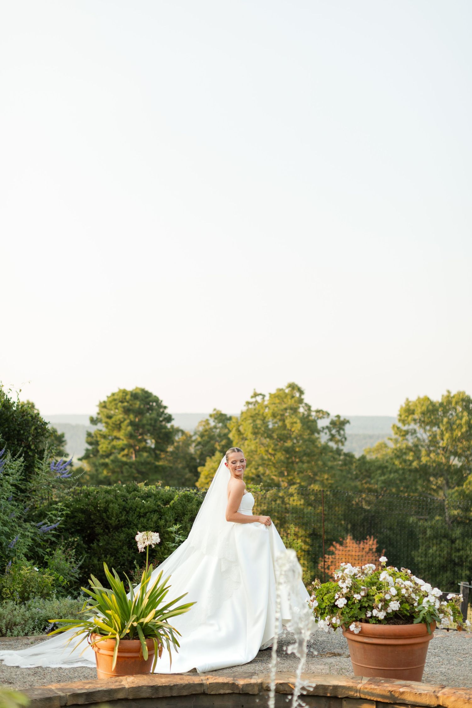 A flowing white wedding dress train cascades across a garden terrace with potted plants and trees in the background.