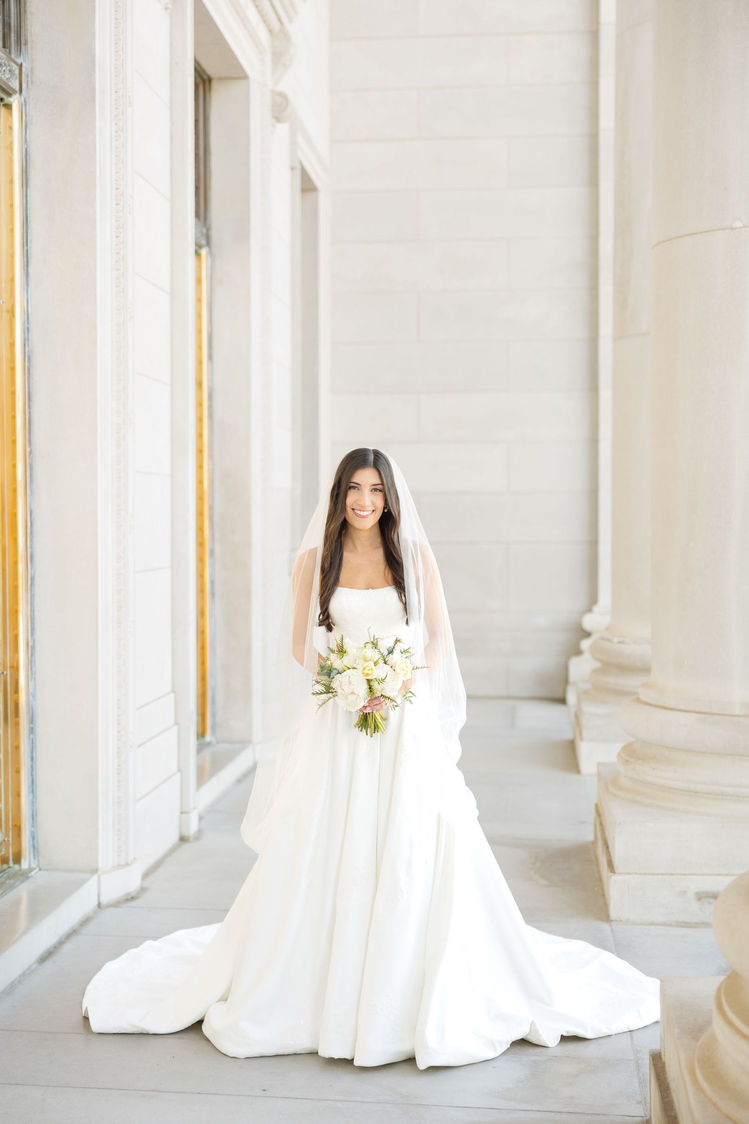 A bride in a classic white gown poses in a bright marble colonnade holding a white floral bouquet.
