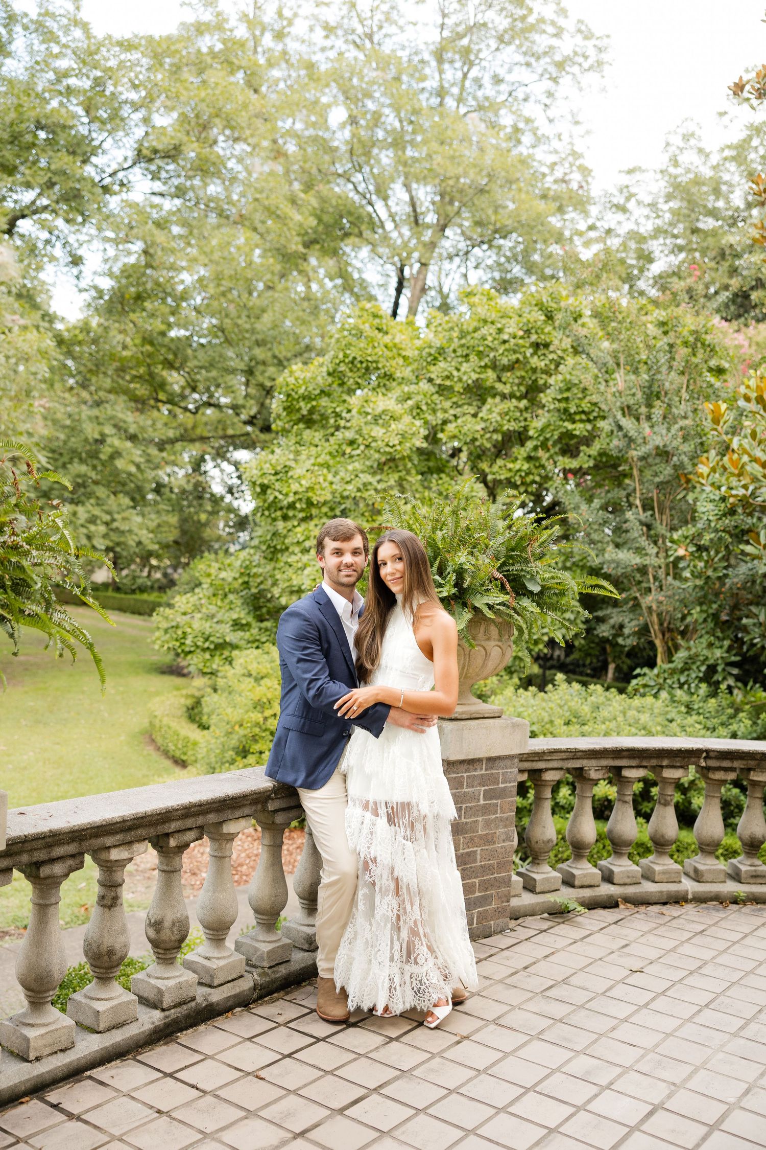 A couple sits together on a stone terrace surrounded by lush greenery and ornate balusters on a sunny day.