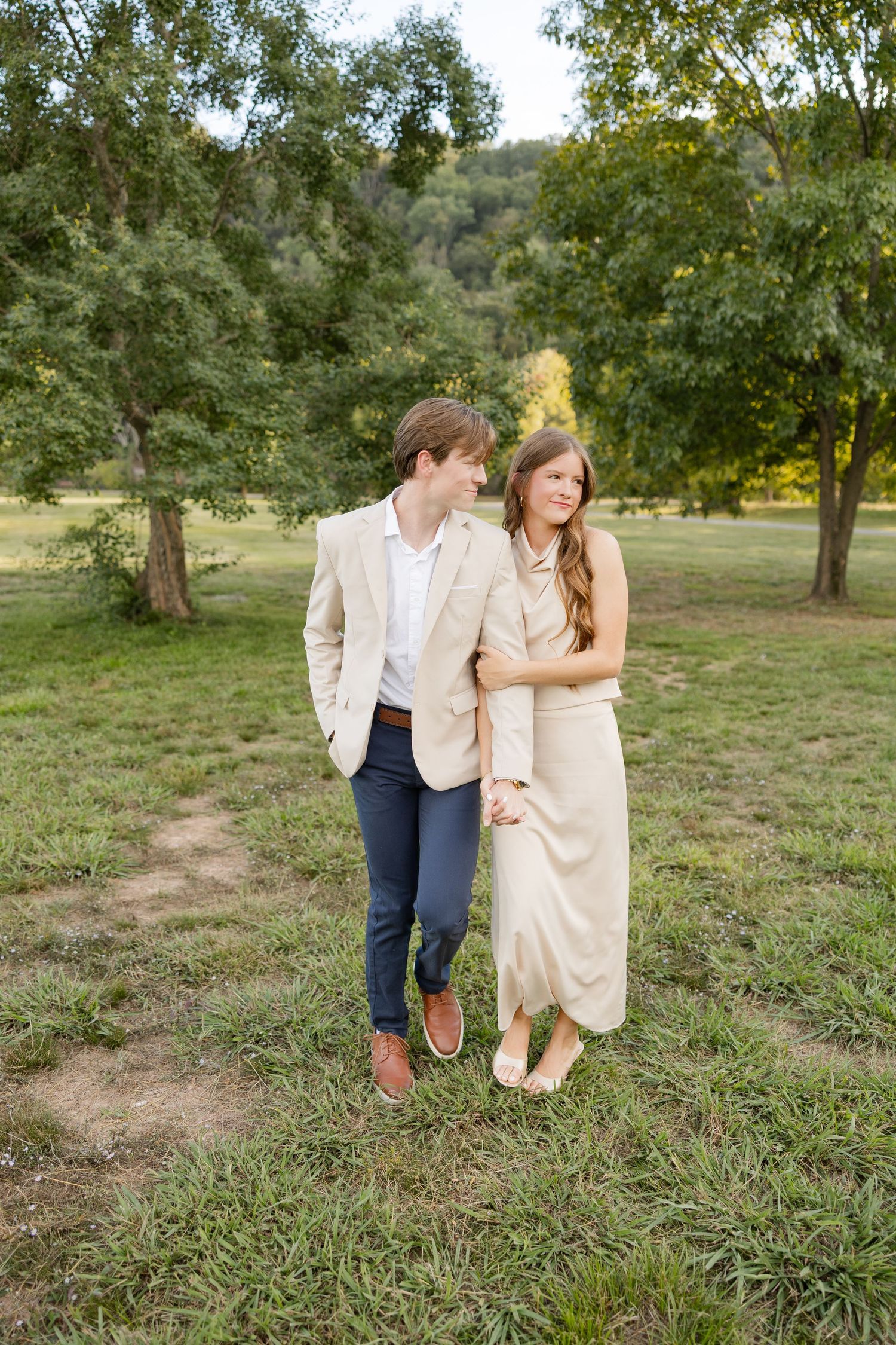 A couple shares a romantic moment walking together through a grassy orchard on a sunny afternoon.