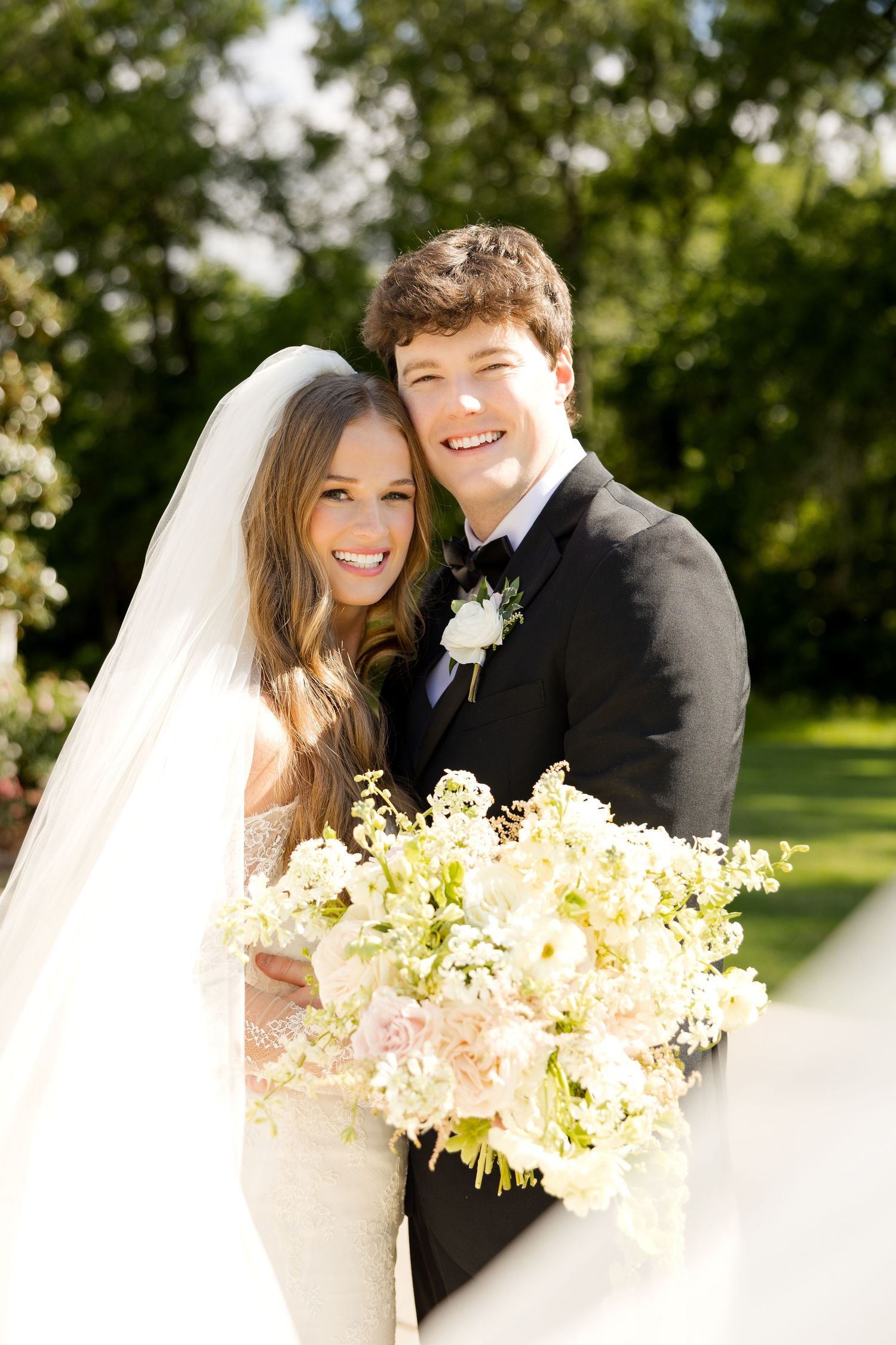 A smiling bride in a white gown and veil holds a lush floral bouquet beside a groom in a black tuxedo outdoors.