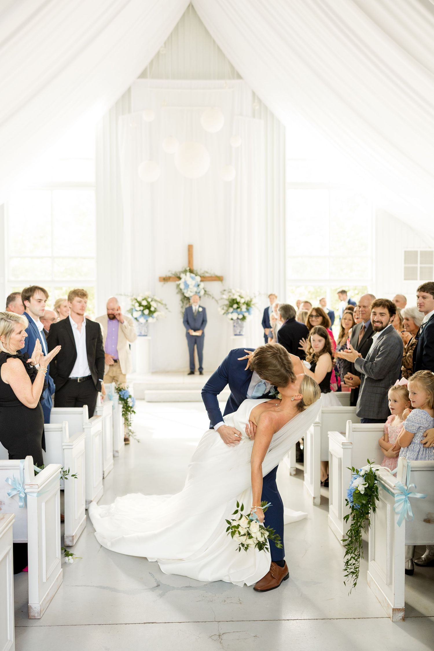 Wedding ceremony in a bright white chapel with guests seated in wooden pews.Cold Springs Events