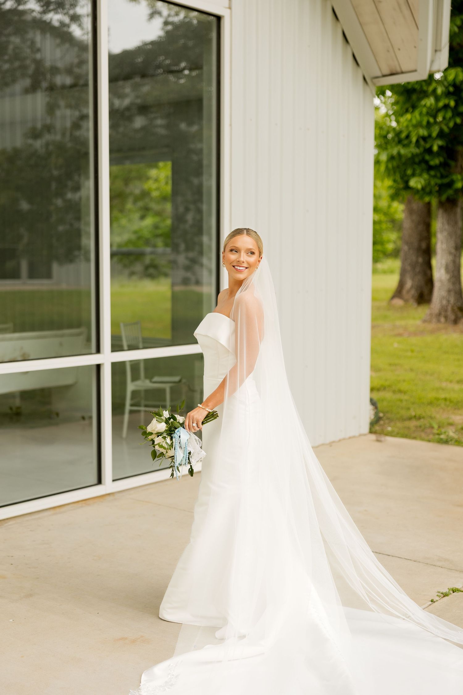 A flowing white wedding dress train and veil trails behind on a covered porch with greenery visible outside.Cold Springs Events.