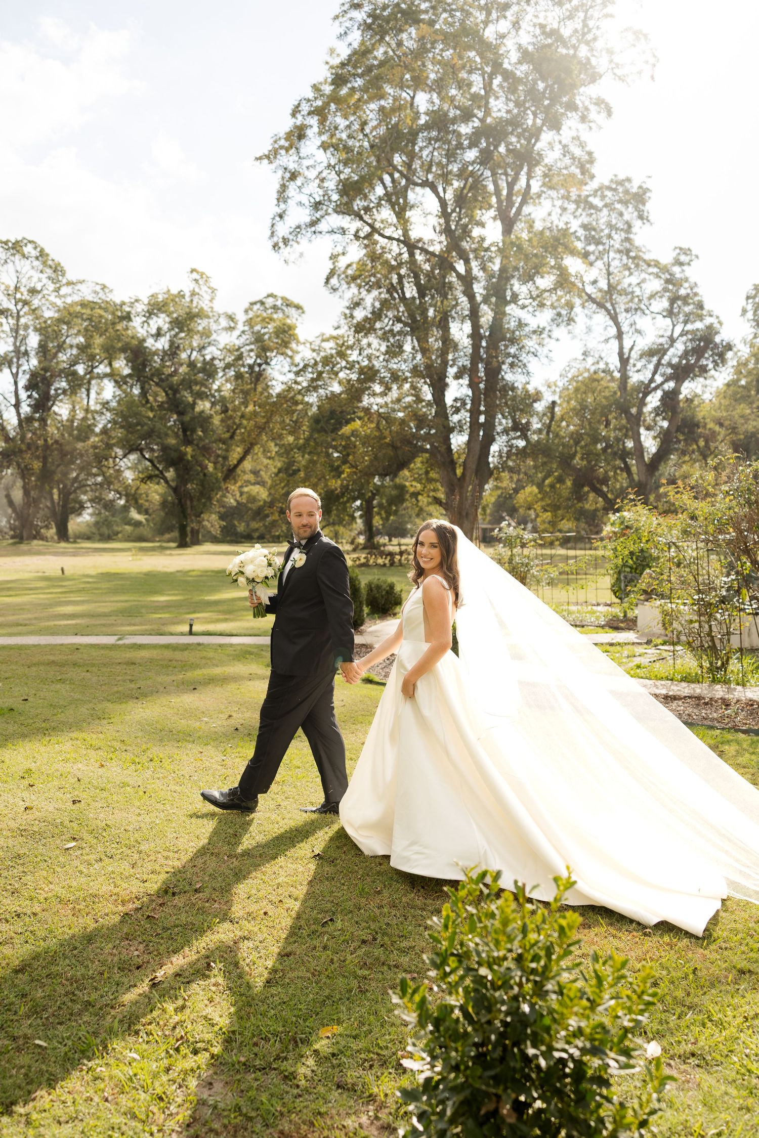 Couple walking together on sunlit lawn with bride's dress trailing behind.Marlgsate Plantation