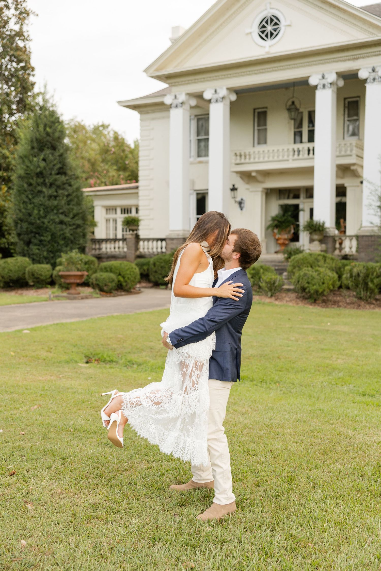Couple in wedding attire sharing a romantic dip pose in front of a white colonial-style building.