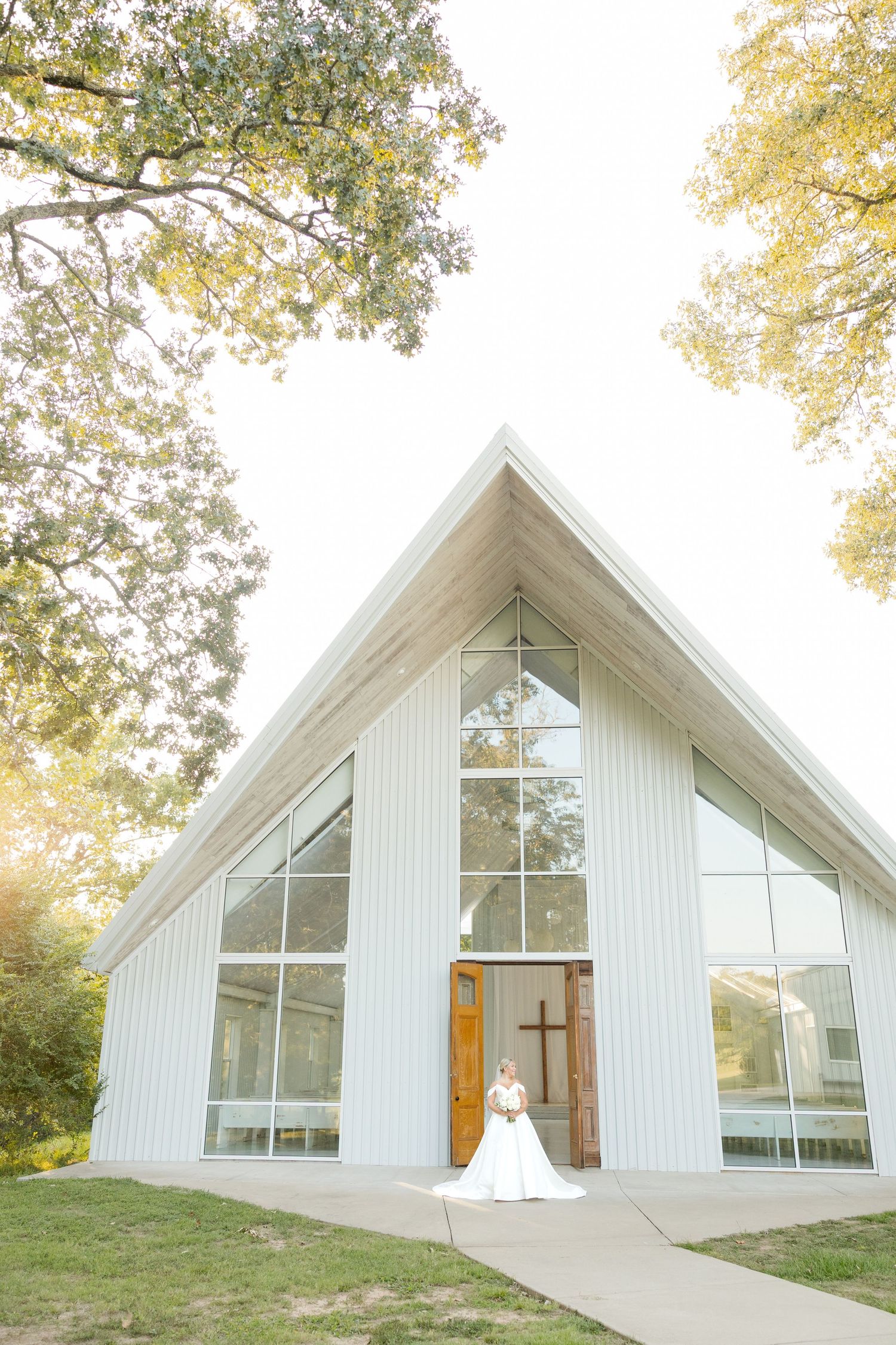 A modern white chapel with large windows and wooden cross framing a bride in her wedding dress.