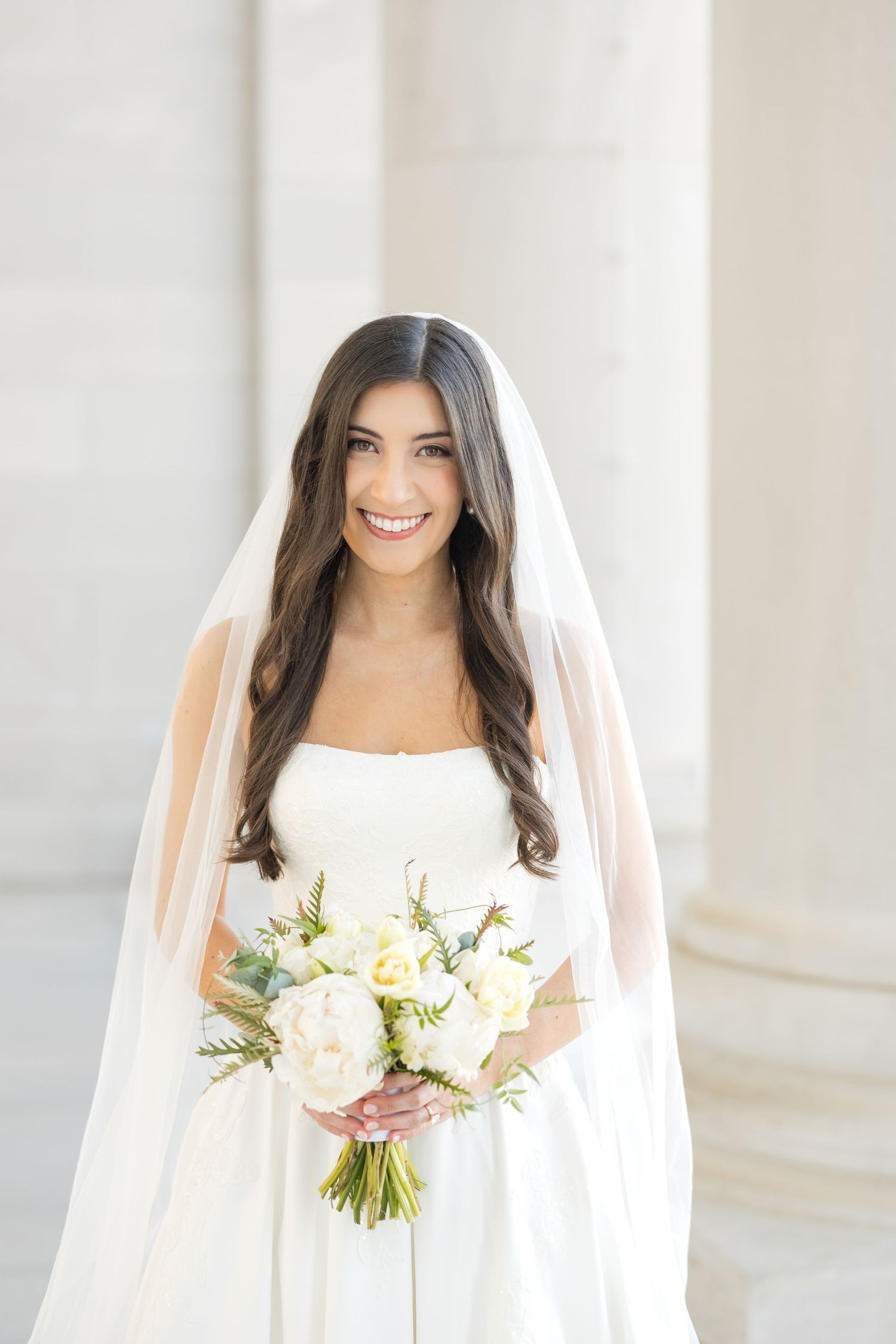 A bride in a strapless white gown holds a fresh bouquet against a bright architectural backdrop.