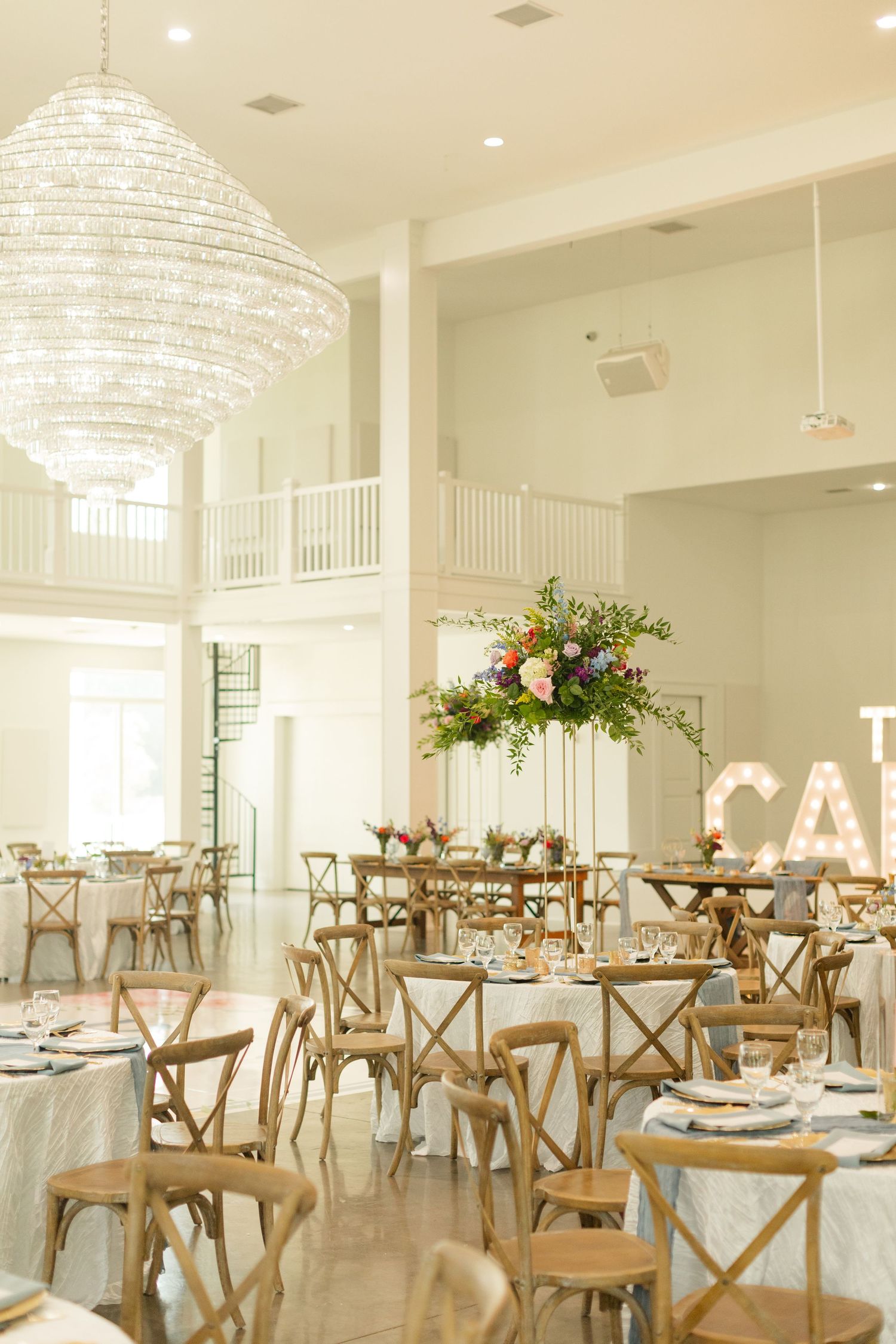 Elegant white wedding reception venue with rustic wooden cross-back chairs and crystal chandeliers creating a bright atmosphere.