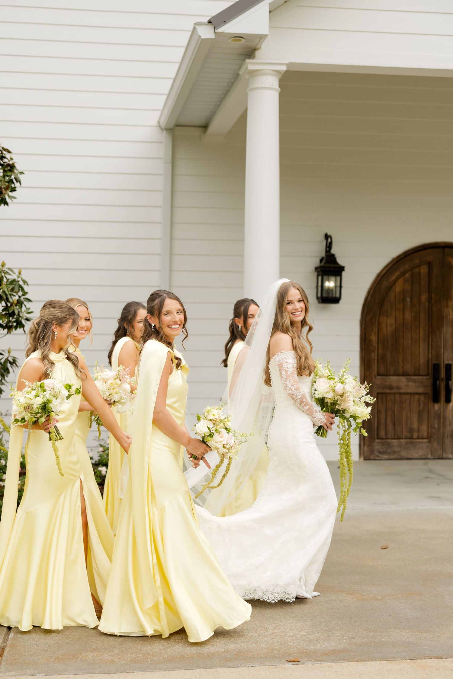 Bride in white lace gown with bridesmaids in yellow dresses holding bouquets outside a white chapel.