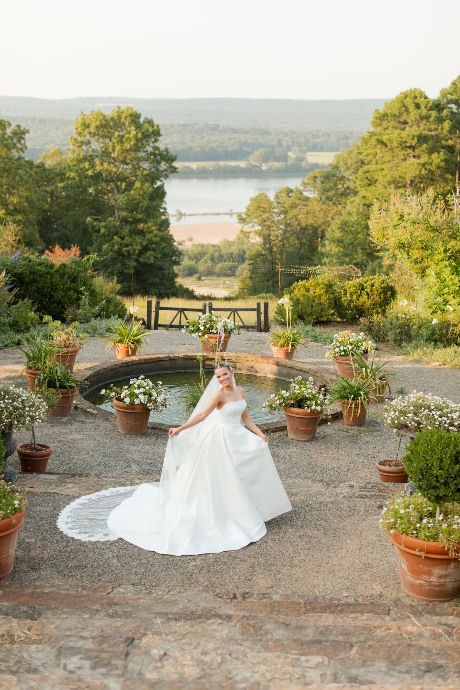 A stunning bridal portrait on stone steps overlooking a lake, surrounded by potted flowers and greenery at sunset.