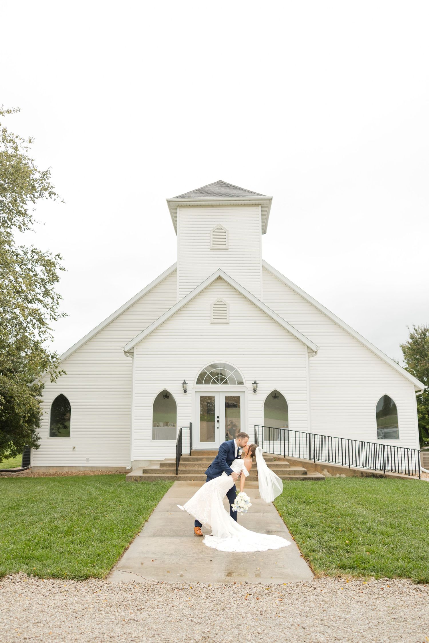 A charming white country church serves as backdrop for elegant wedding portraits on a grassy hill.