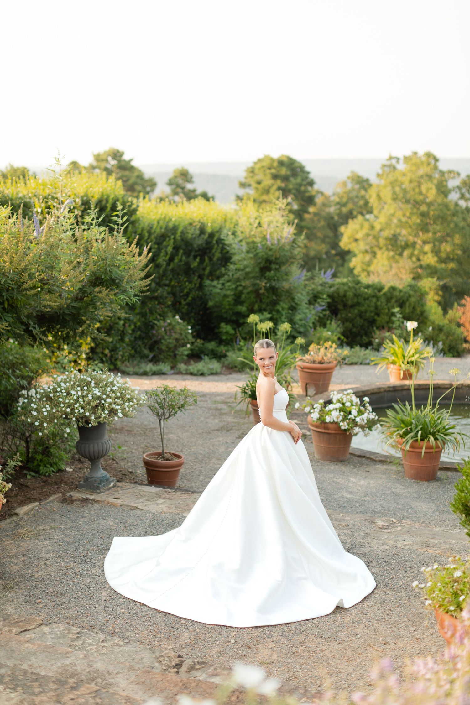 A stunning white wedding dress with a long train displayed in a garden setting with terracotta pots and greenery.
