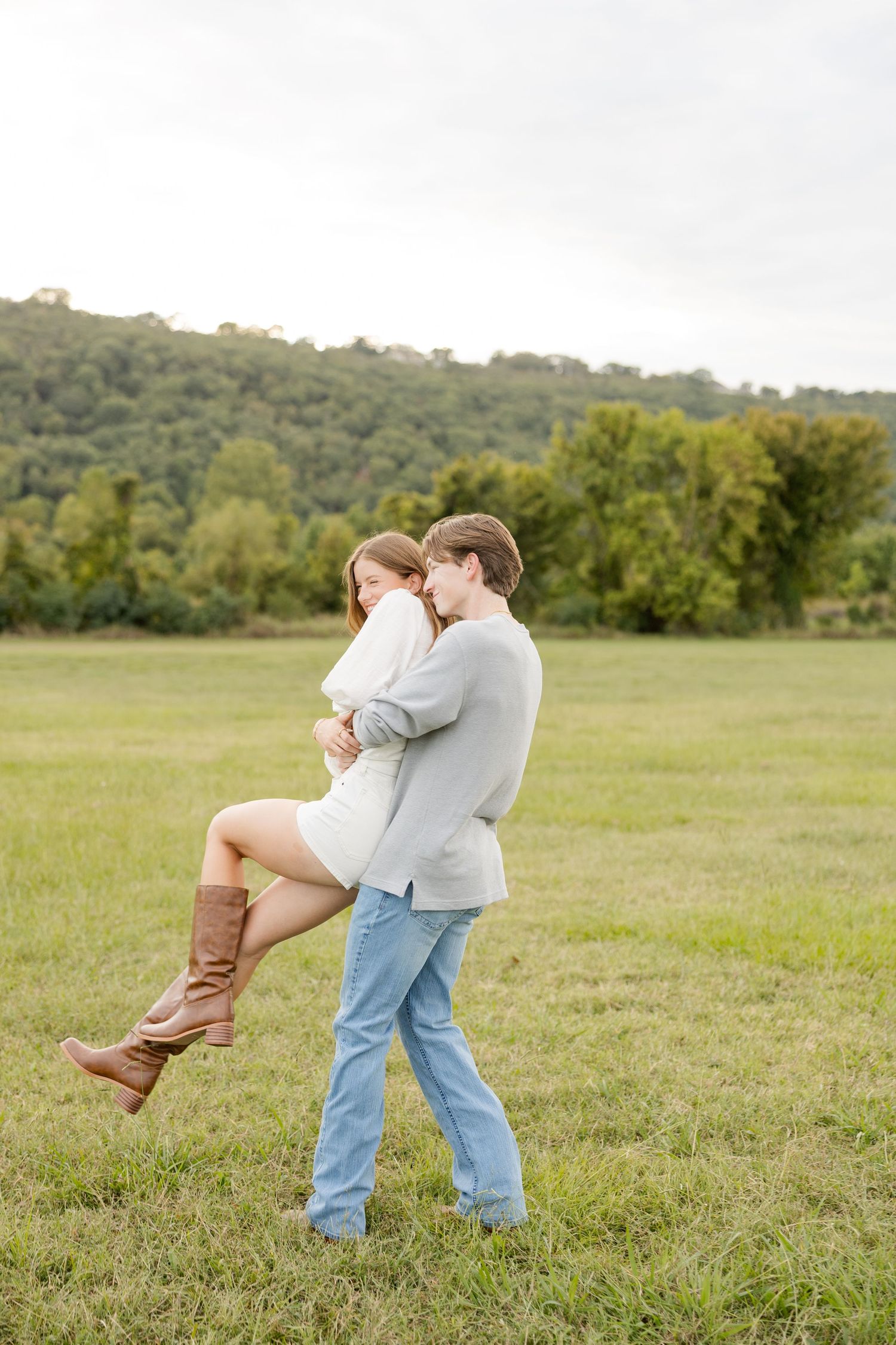 A couple embraces playfully in a grassy field with trees in the background on an overcast day.