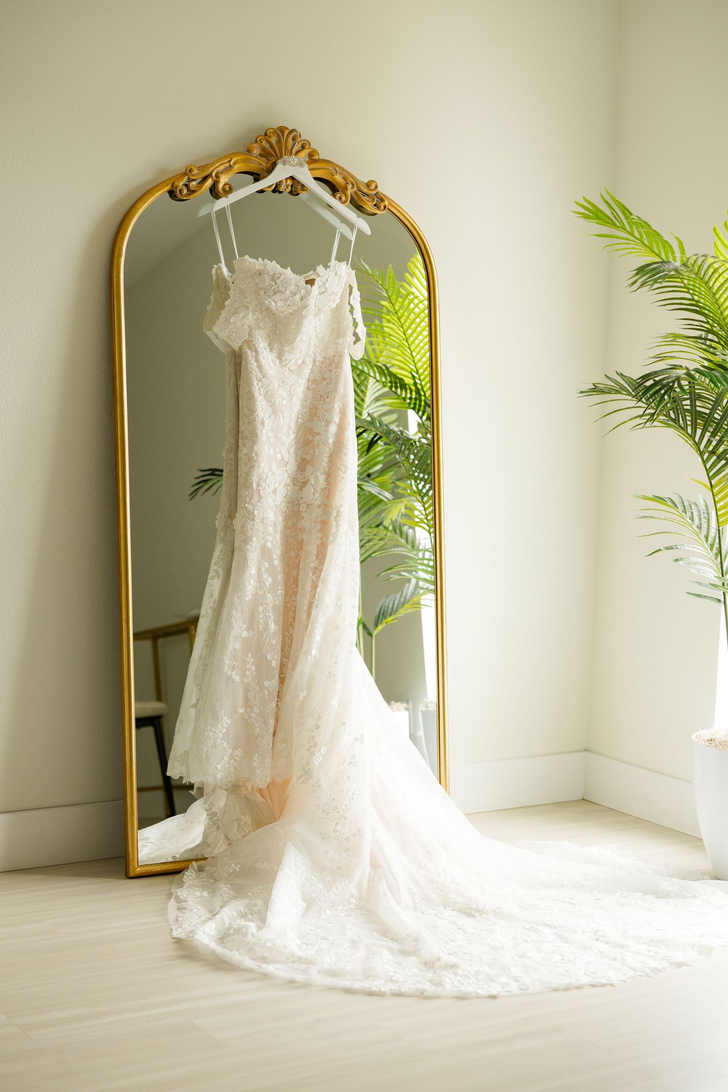 Elegant white lace wedding dress hanging on ornate gold mirror with palm plants in bright corner room.