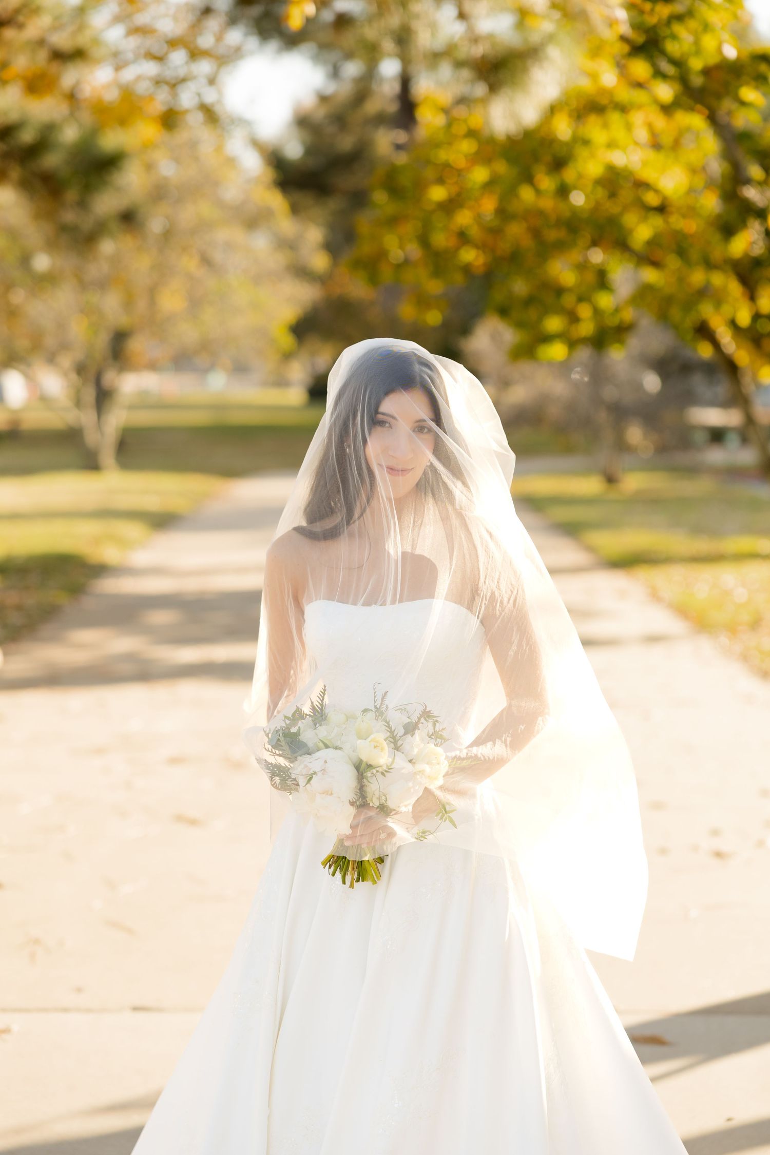 A bride in a white wedding gown and veil holds a bouquet of white flowers on a sunny autumn day in a park.