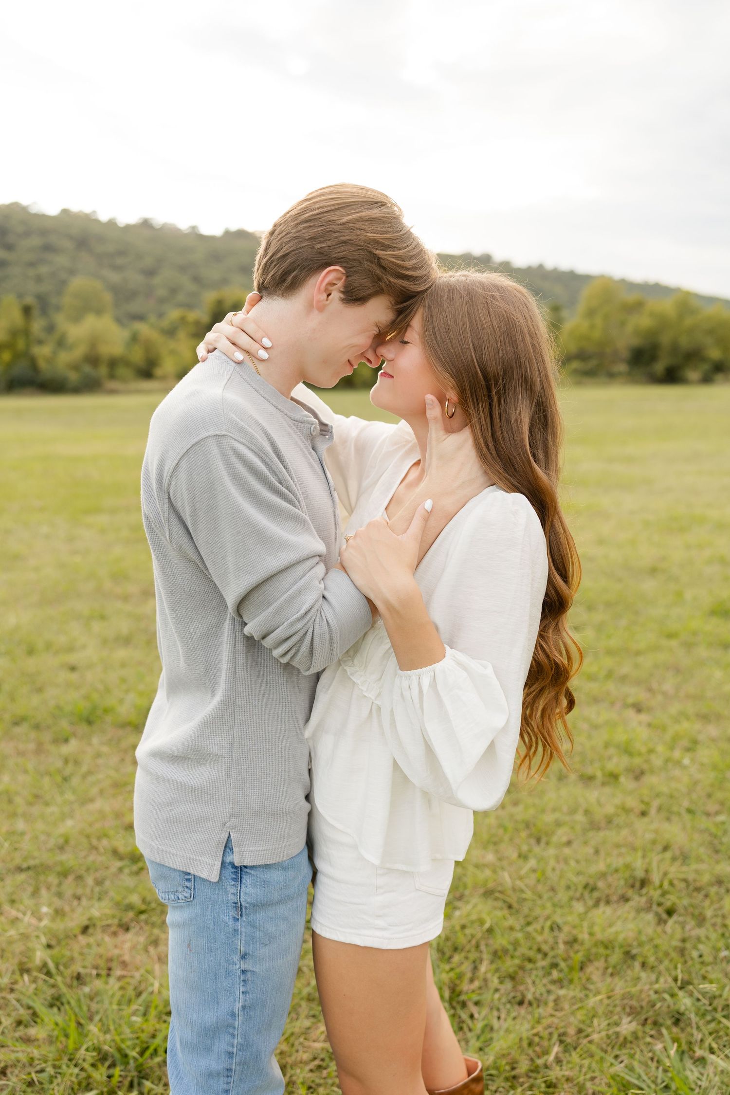 A romantic couple embraces in a grassy field with rolling hills and trees in the background.
