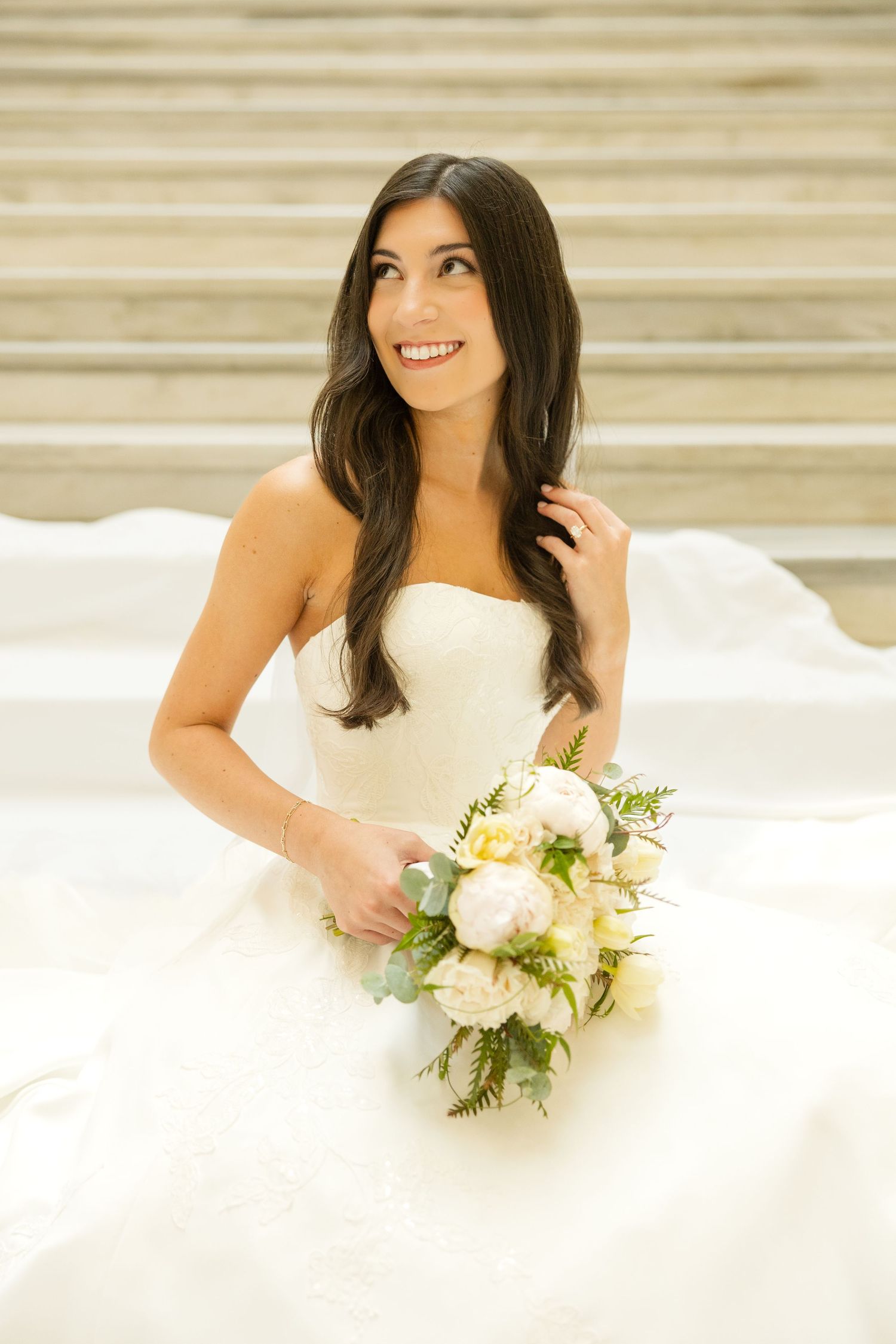 Bride in strapless gown holds white floral bouquet while smiling.