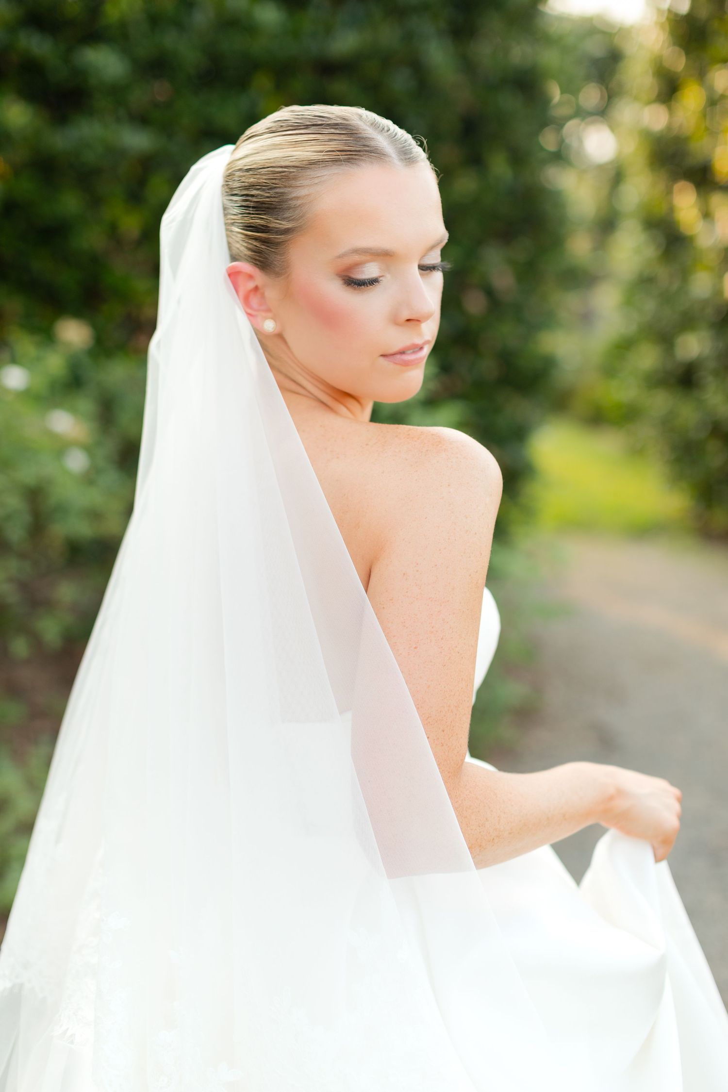 A bride in a flowing white veil poses elegantly against a sunlit garden background.
