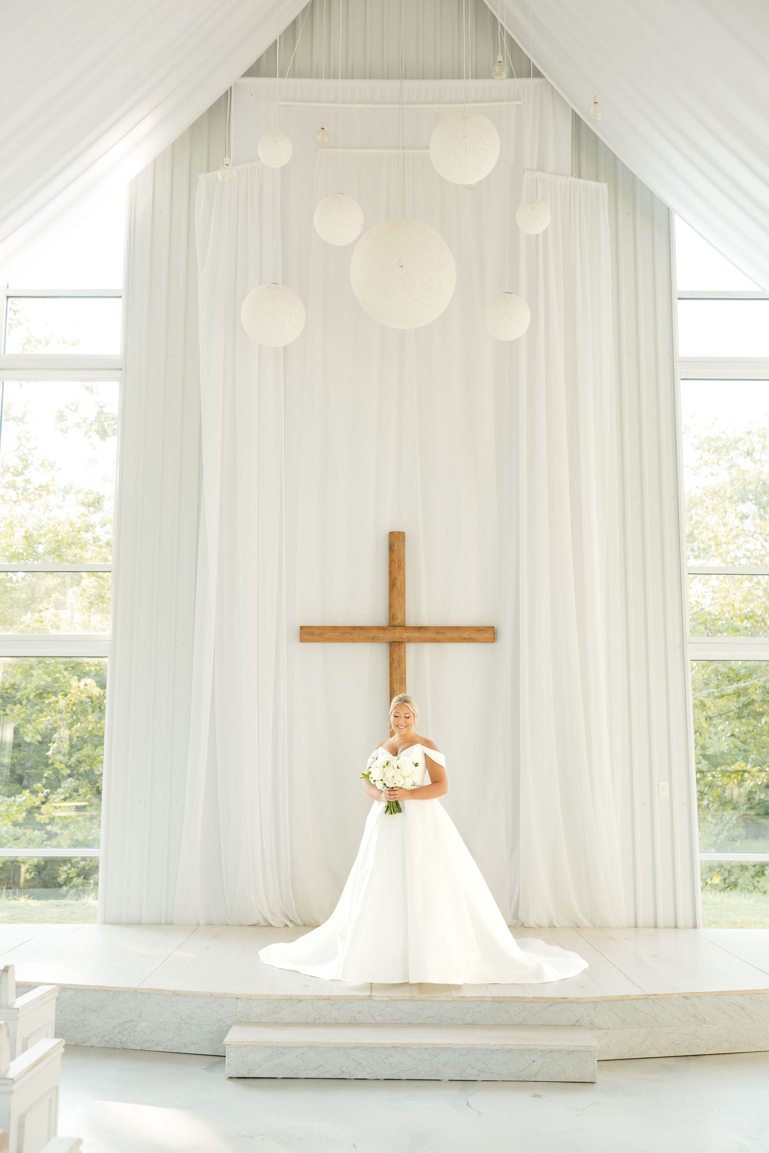 A stunning white chapel interior with flowing sheer curtains, wooden cross, and natural light streaming through windows.