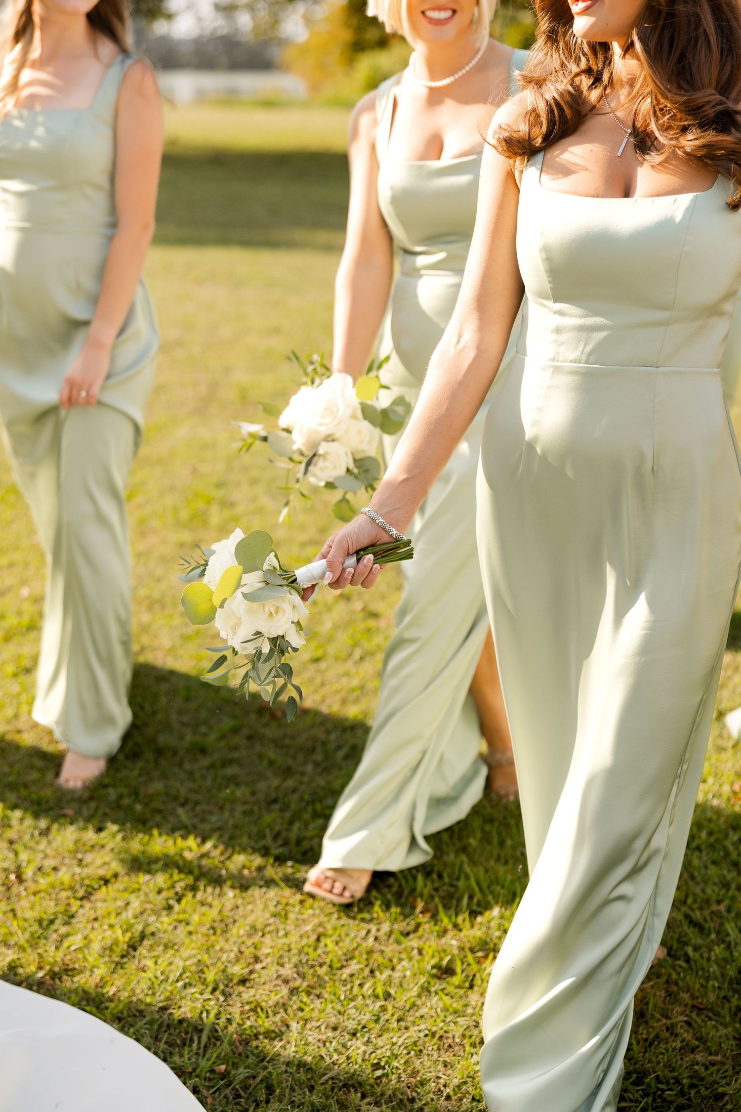 Bridesmaids in matching sage green jumpsuits holding white bouquets pose on a sunny lawn.