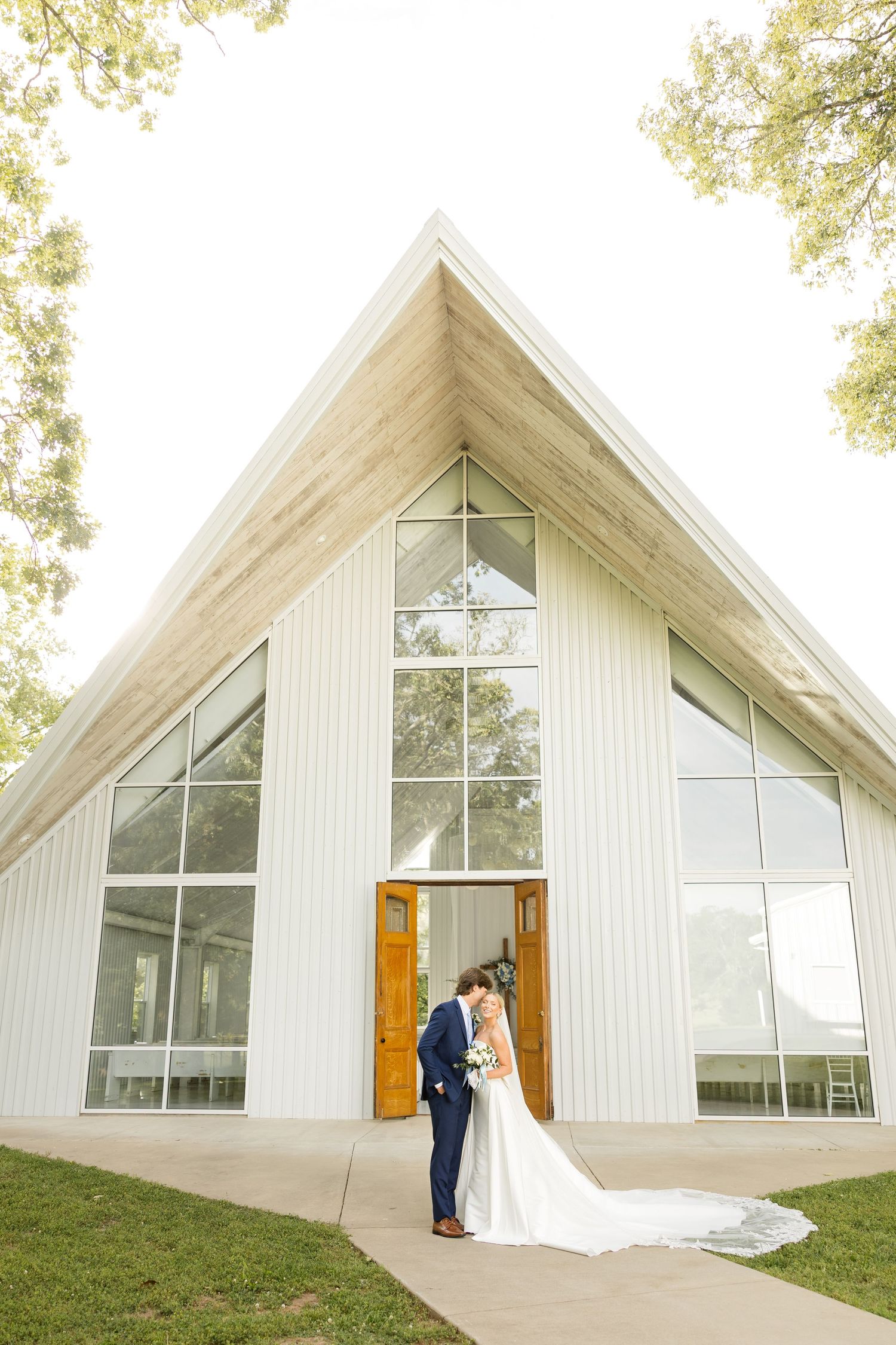Modern white chapel with large windows and wooden doors set against green trees and grass.Cold Springs Events