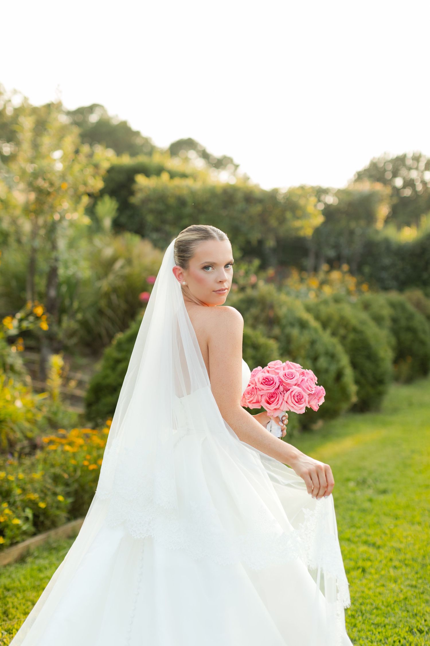 A bride in a flowing white wedding gown poses gracefully in a lush garden setting during golden hour.