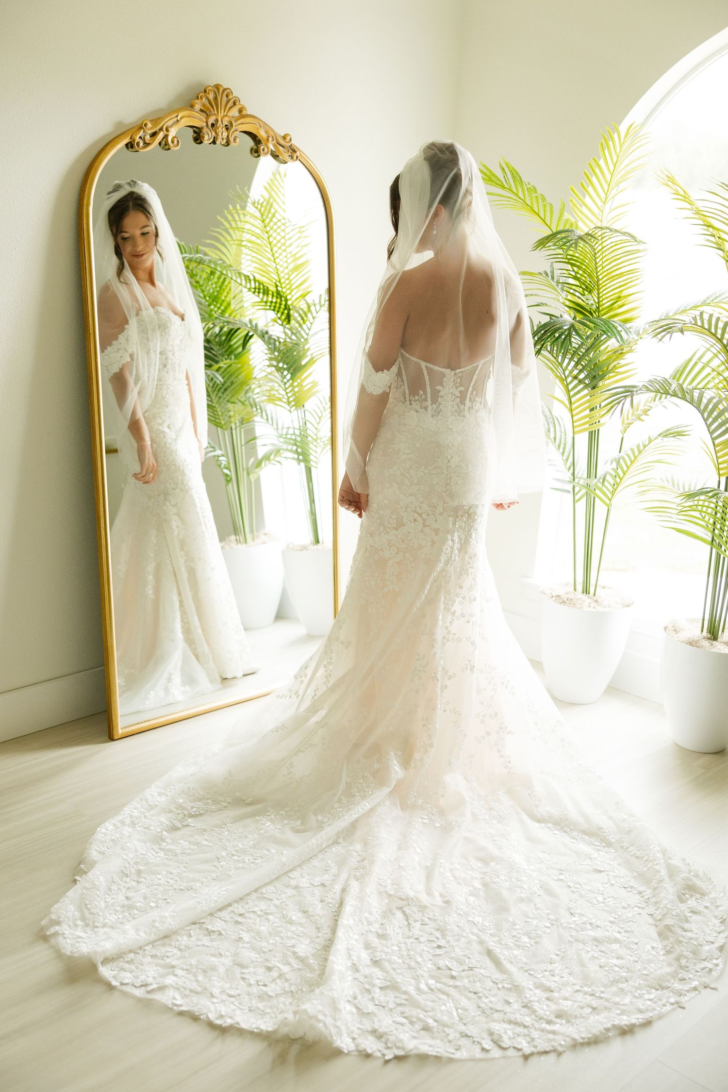 An elegant wedding dress with lace details and long train displayed in a bright room with palm plants and gold mirror.