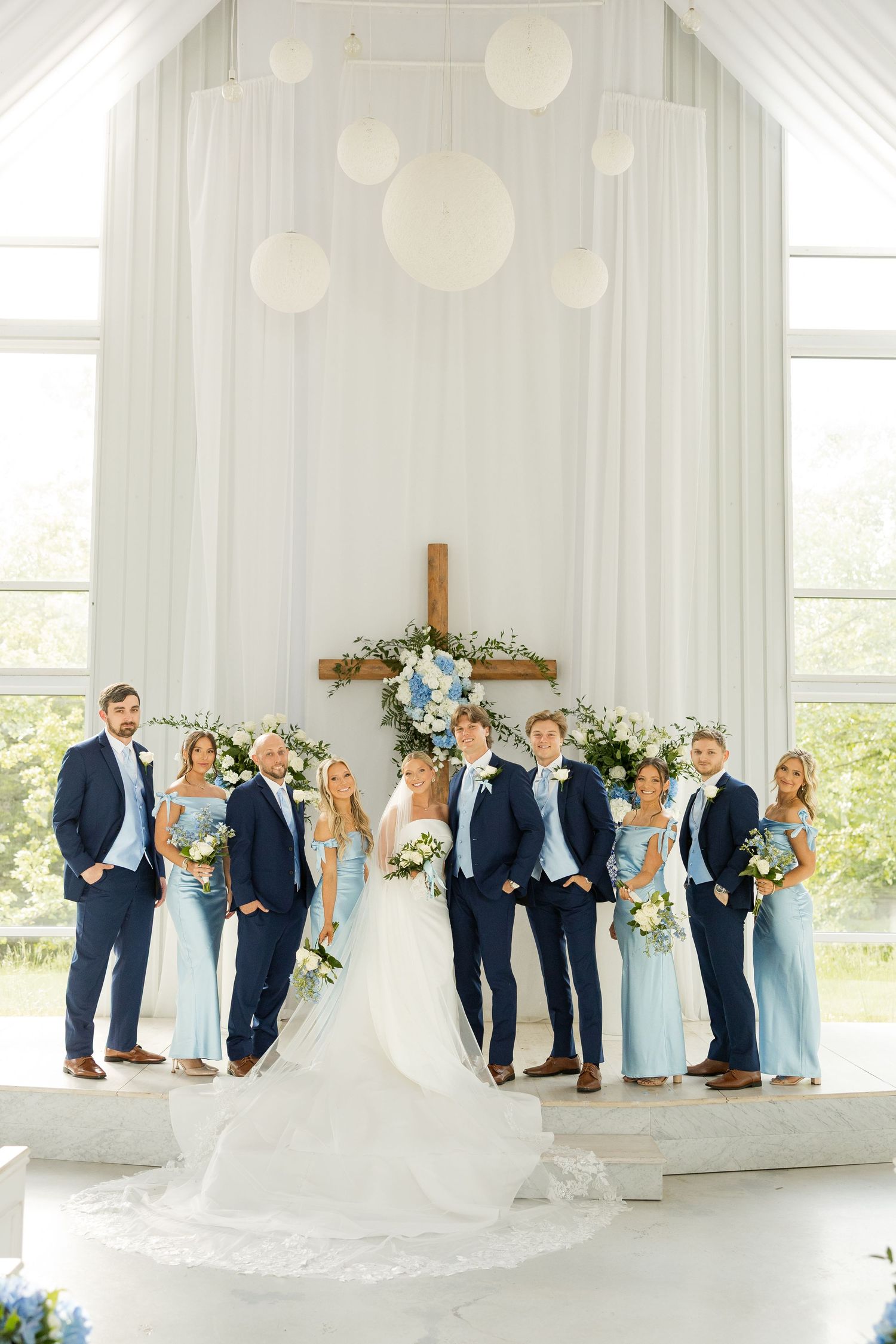 Wedding party in navy suits and light blue dresses poses in front of a white cross altar.Cold Springs Events.