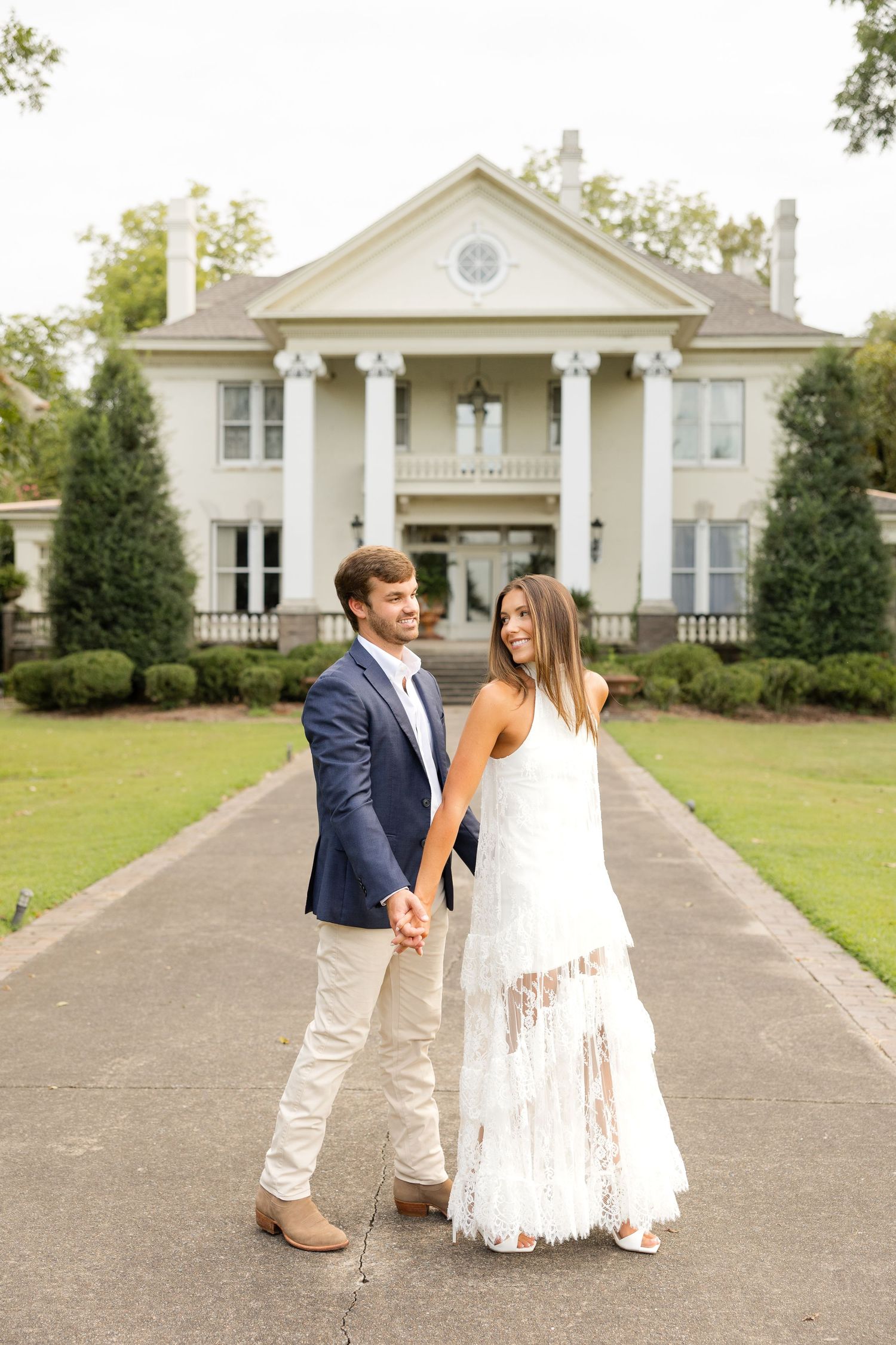 A couple in formal attire shares a romantic moment on a driveway with a white house in the background.