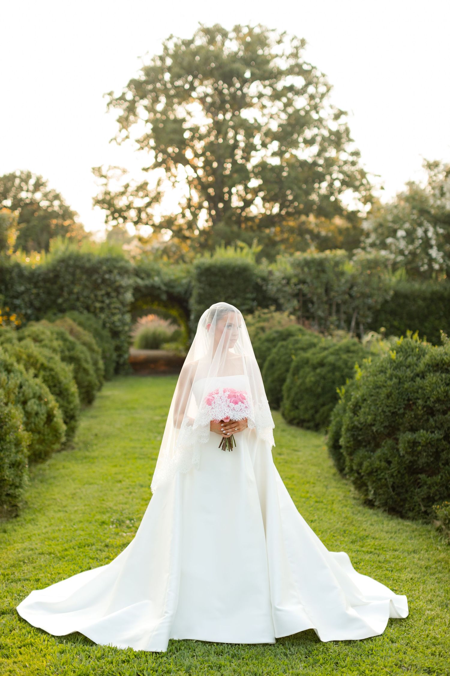 A wedding dress with long train spreads across lush green grass with manicured hedges in the background.