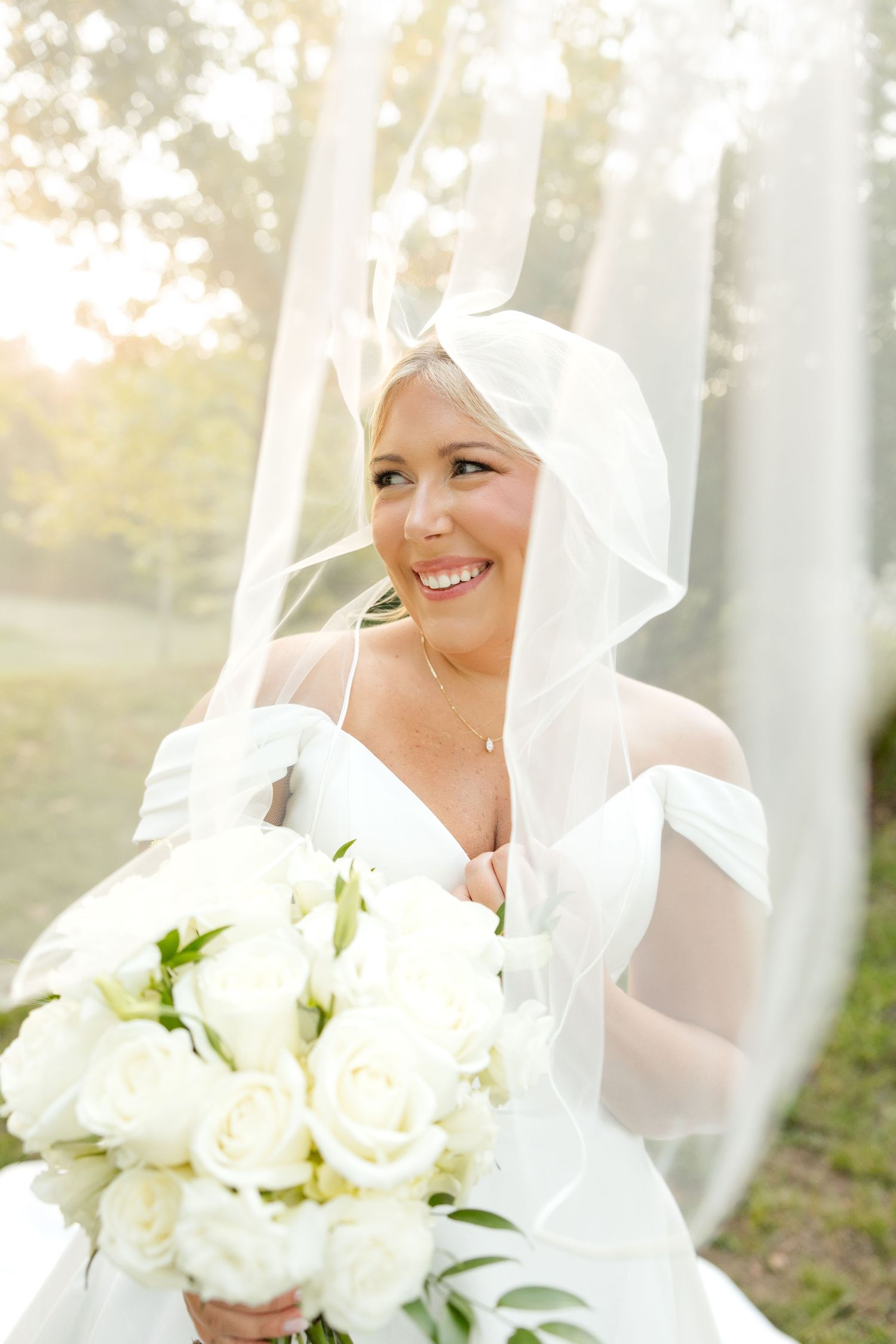 A joyful bride in a white veil holds a bouquet of white roses on her wedding day in soft natural lighting.