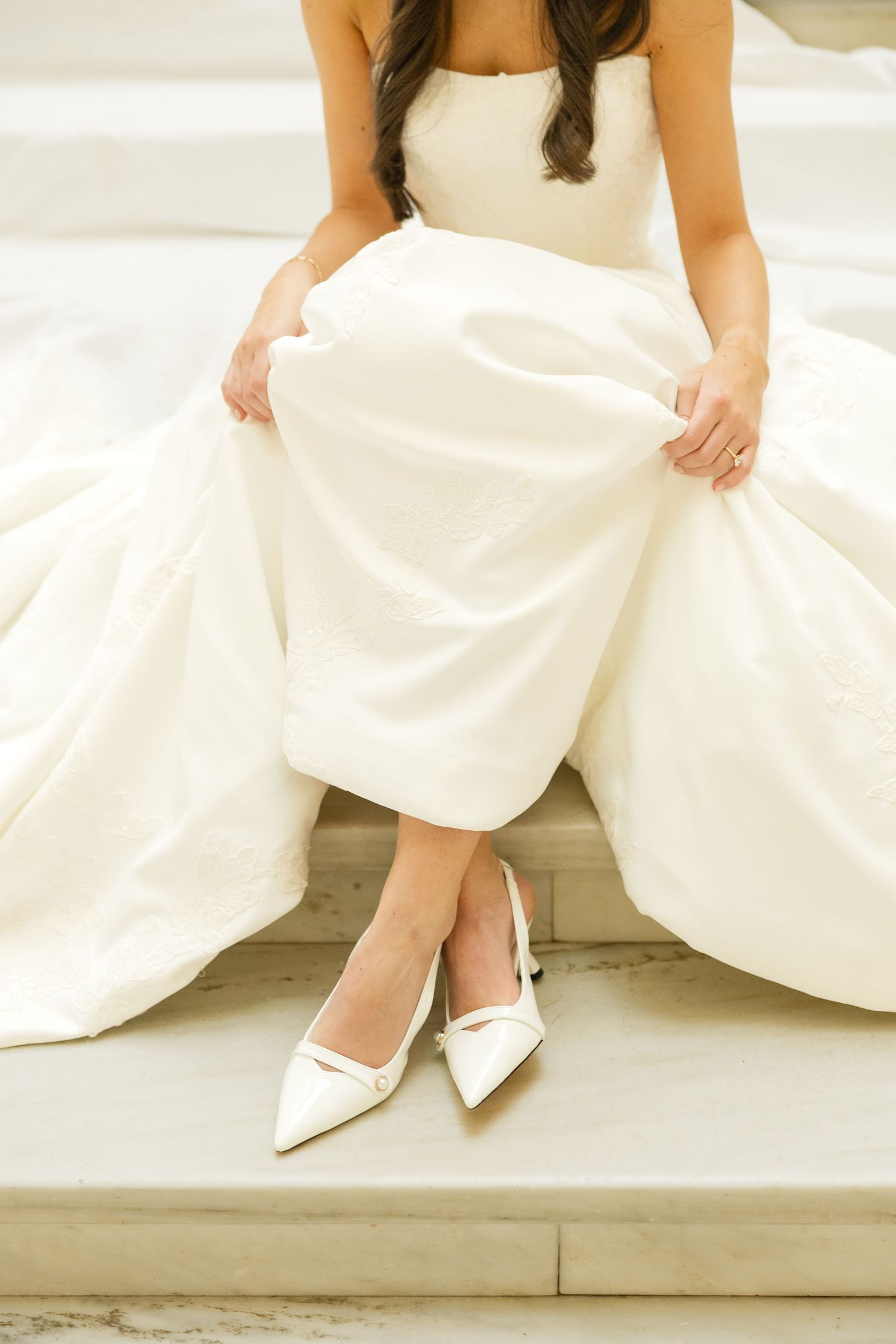 Close-up details of white bridal shoes and dress hem while sitting on marble steps.