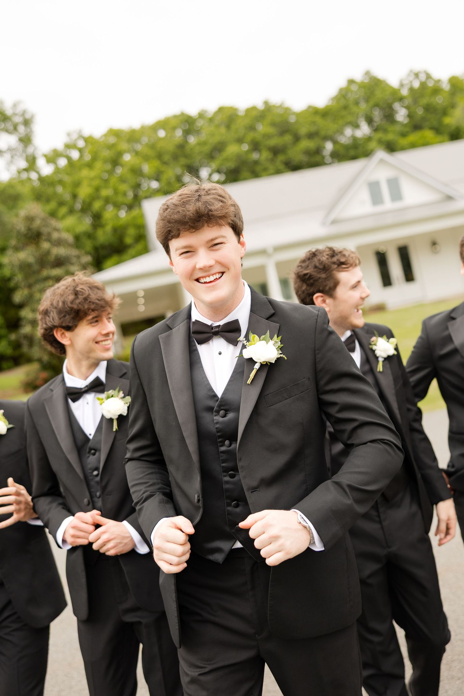 Groom in black tuxedo with bow tie and boutonniere laughs with groomsmen outside white house venue.