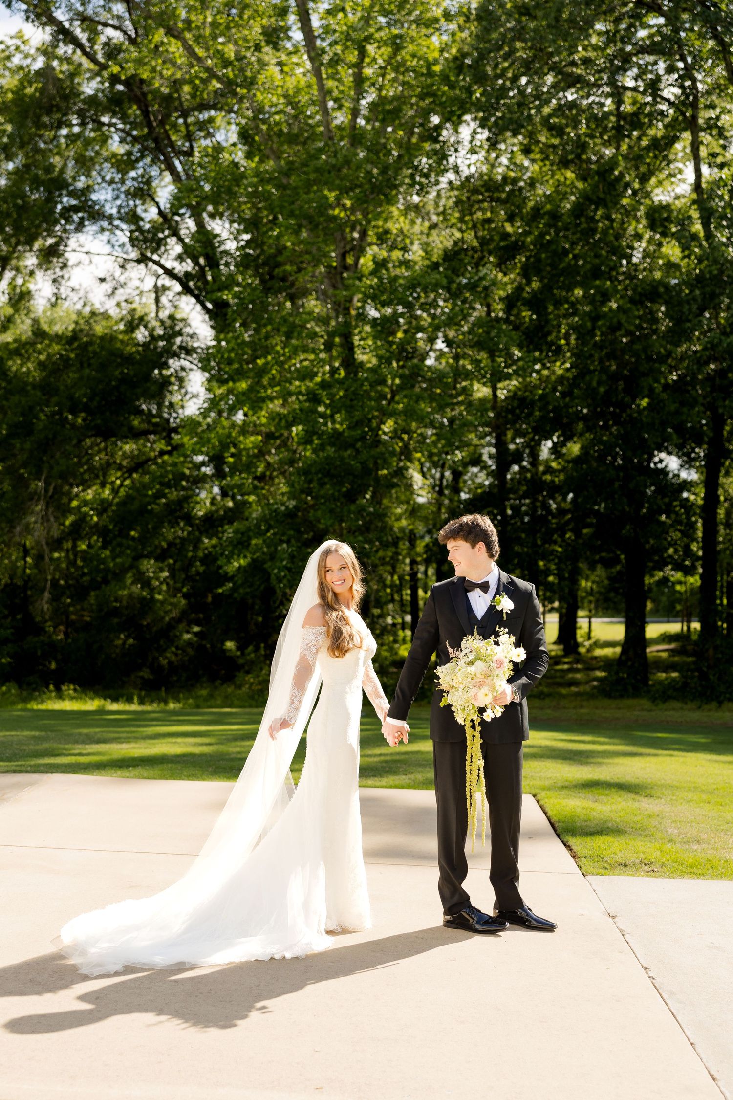 Bride in flowing white gown with long veil and groom in black tuxedo holding hands outdoors.