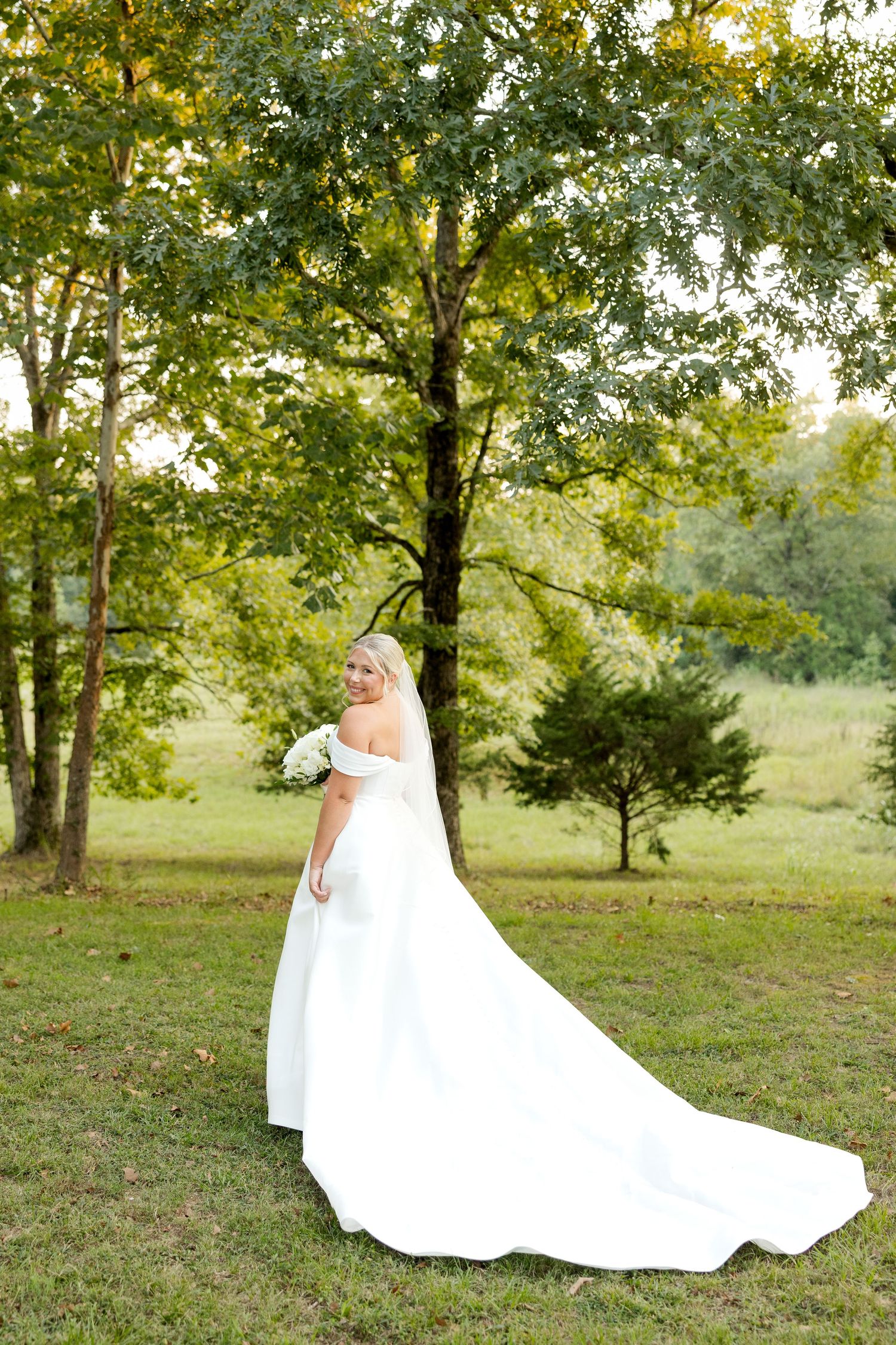 A flowing white wedding dress moves gracefully against a backdrop of lush green trees and grass.
