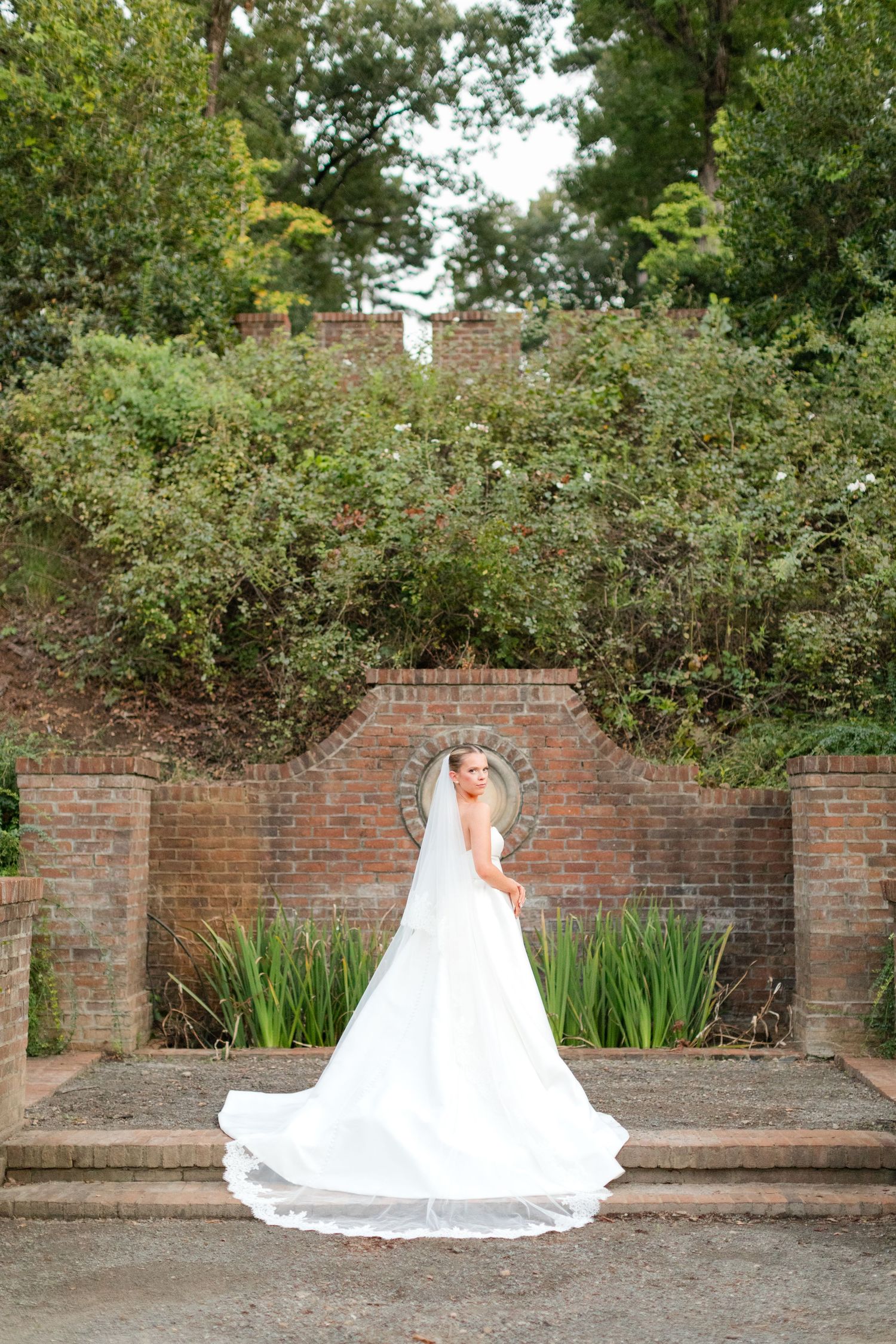 Dramatic bridal portraits against a rustic brick wall with garden foliage and flowing white wedding gown.