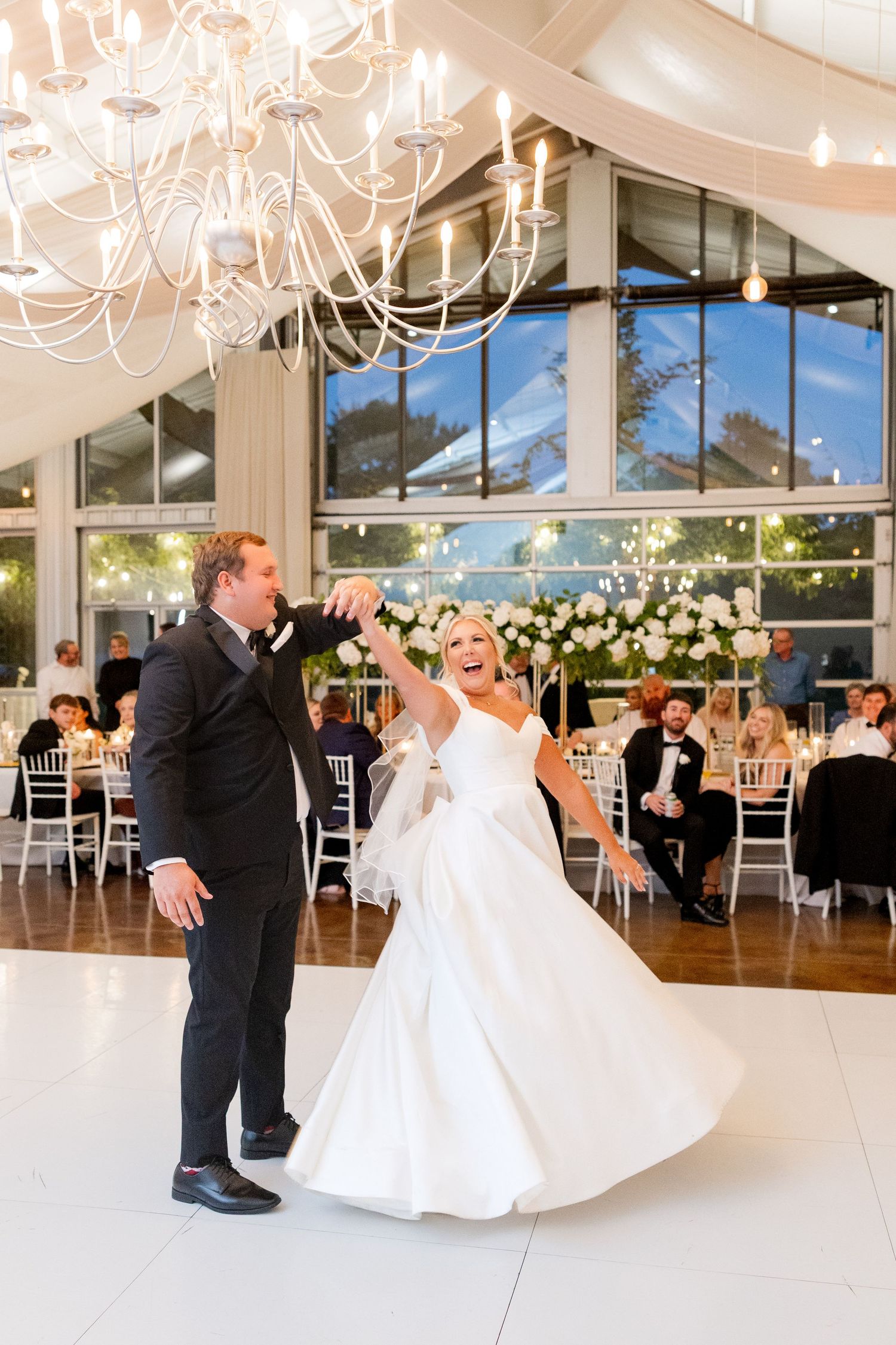 An elegant white wedding reception venue with chandeliers and floor-to-ceiling windows showing couples dancing. Cold Springs Events.