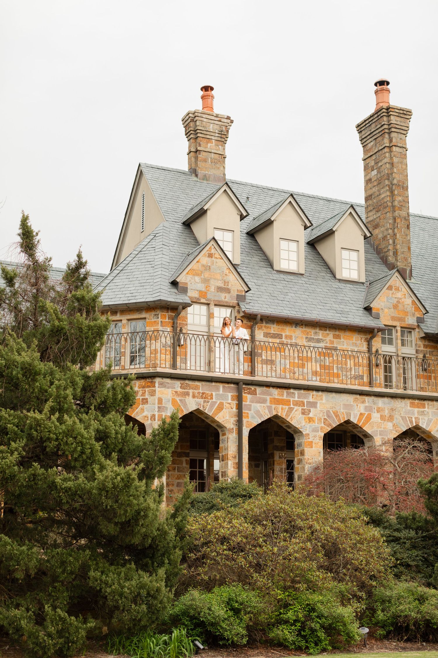 Stone mansion with dormer windows and chimneys sits nestled among evergreen trees and shrubs on a hillside estate. Goodwin Manor.