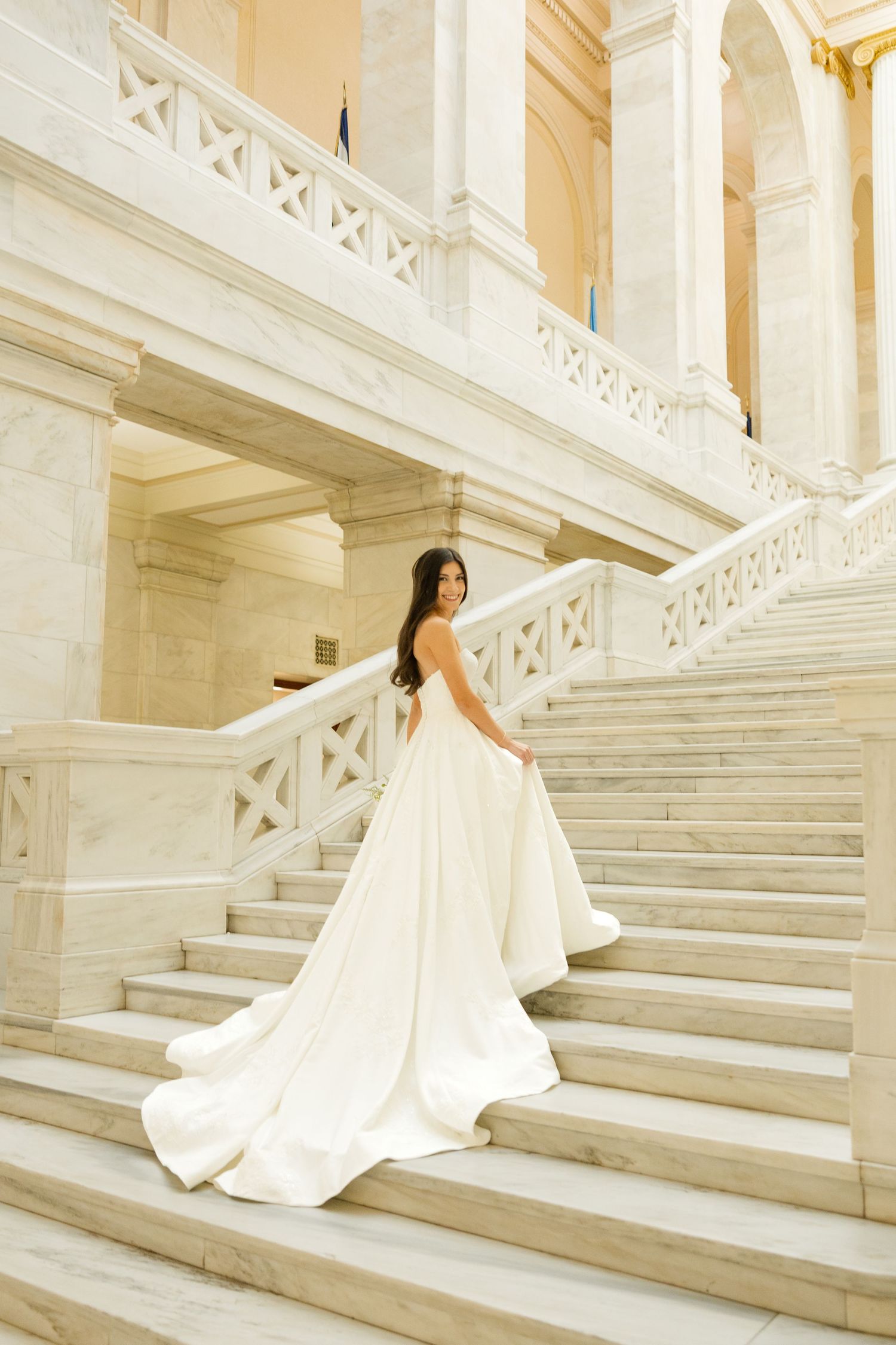 A bride in an elegant white gown poses on ornate marble stairs in a classical building.