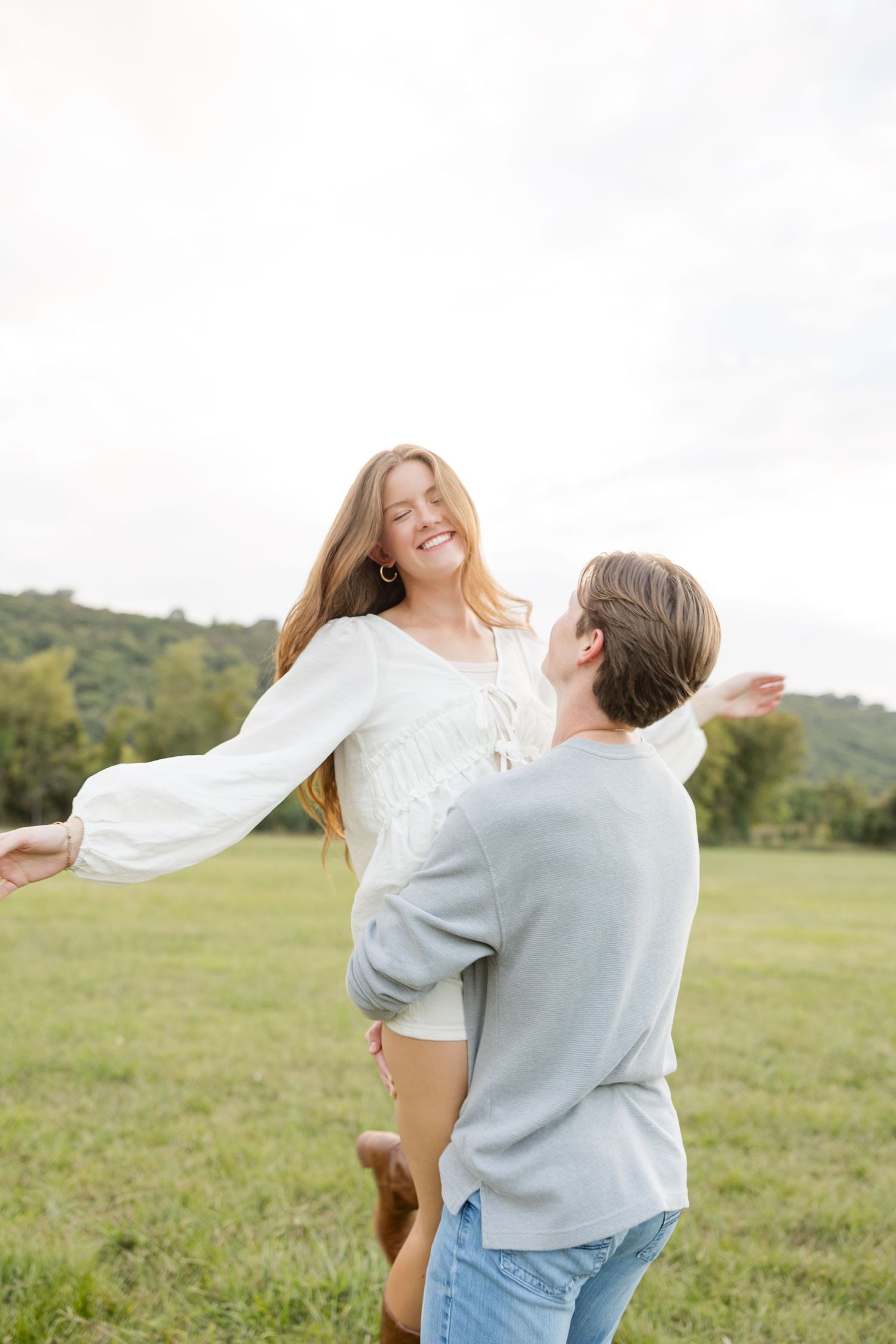 A couple dances and spins joyfully together in a sunny green meadow with rolling hills in the background.