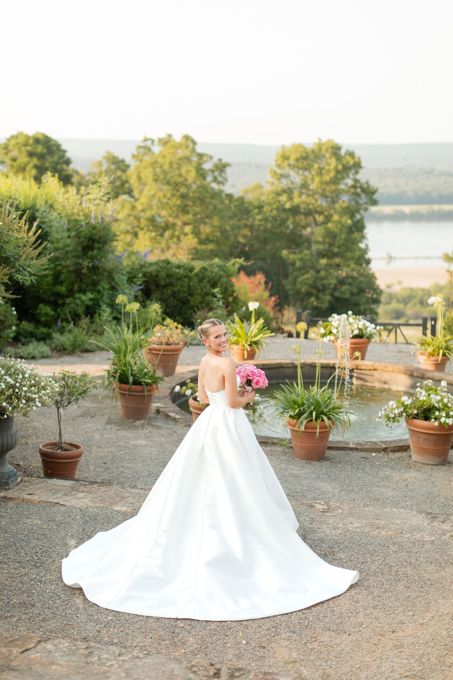 A stunning white wedding gown with a long train displayed in a garden setting with potted plants at sunset.