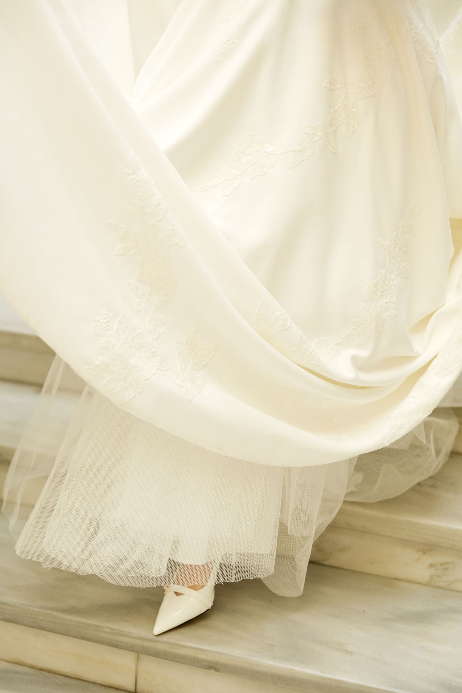 The hem of a white wedding dress sweeps across a light marble floor, showing intricate fabric details.