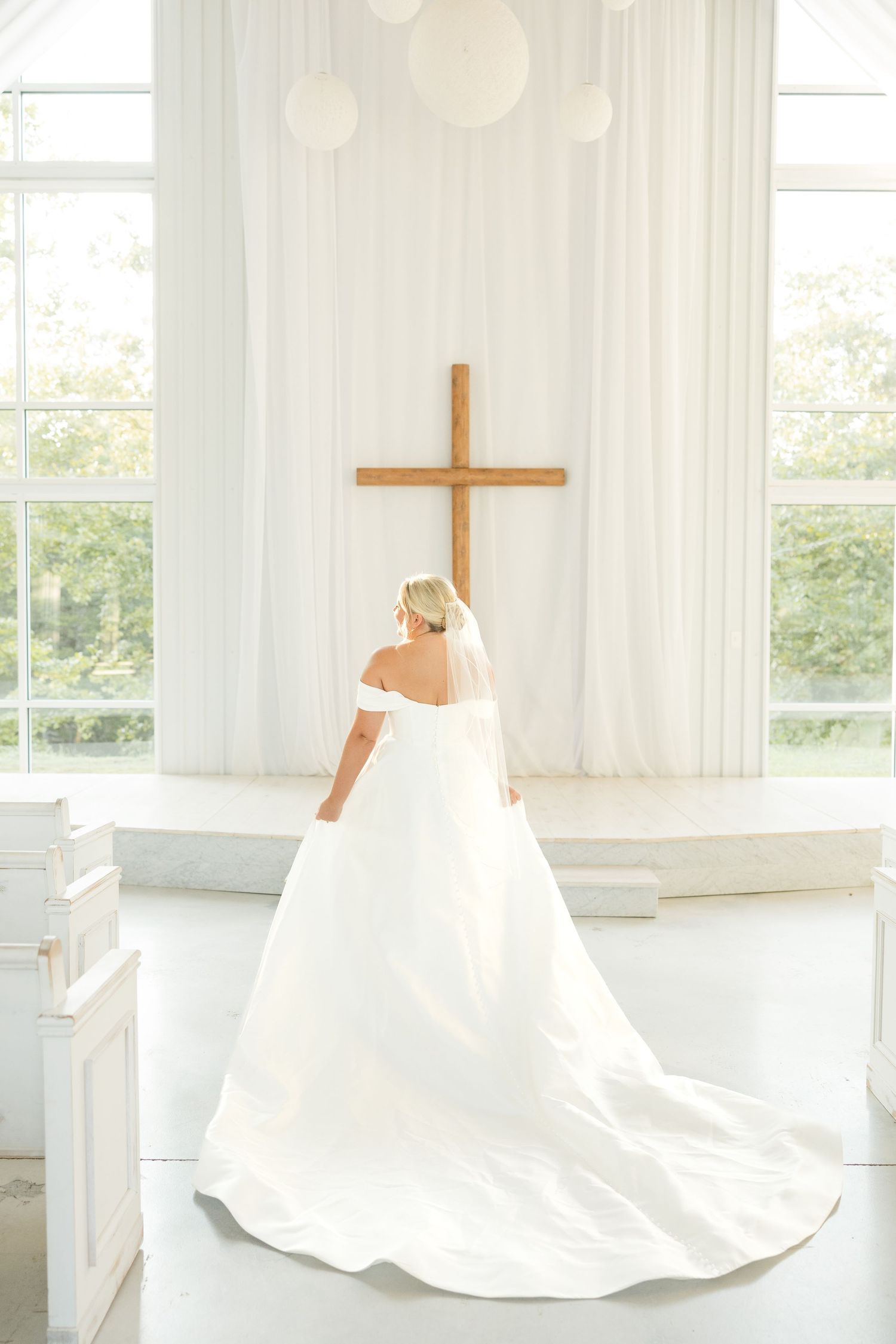 A bride in a white off-shoulder gown stands before a wooden cross in a bright chapel interior.