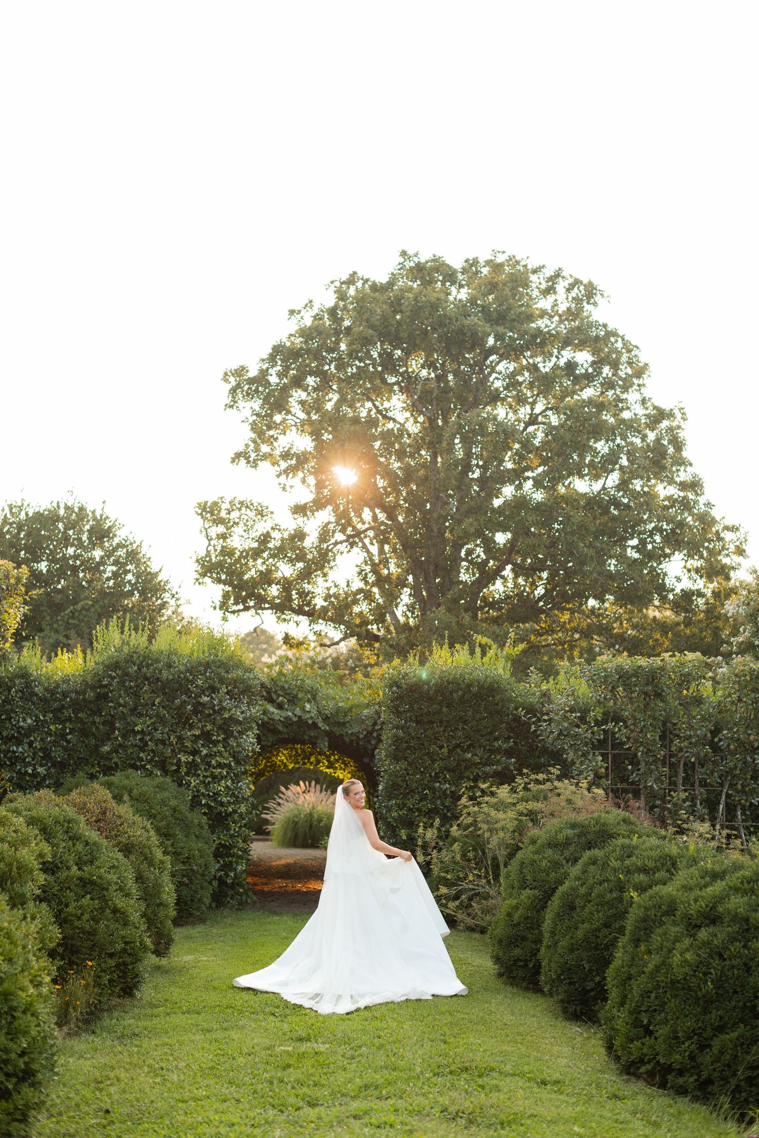 A stunning wedding dress flows across a lush green garden setting during golden hour sunset.